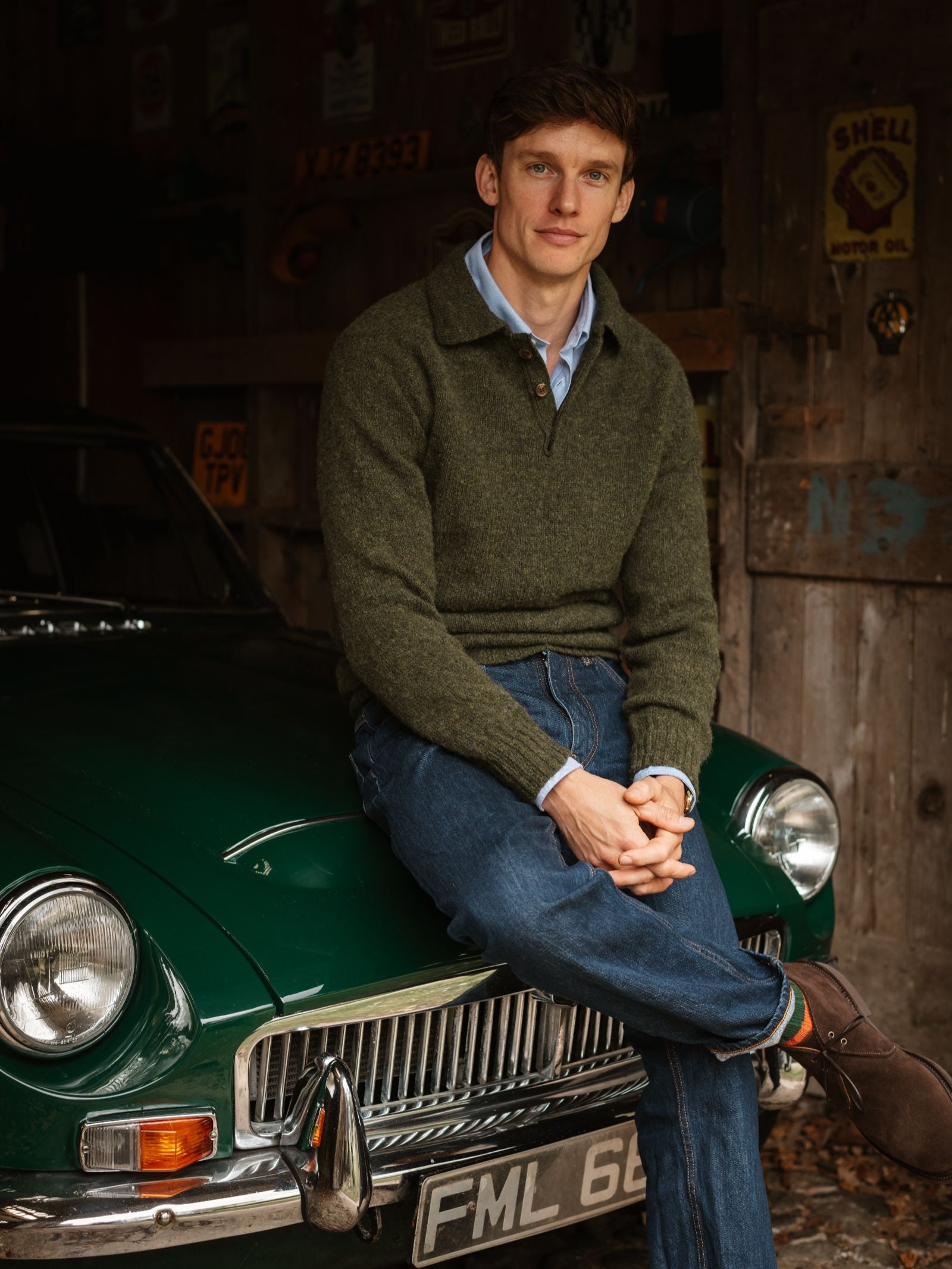A man in a green Campbell's of Beauly Brushed Shetland Rugby Jumper and blue jeans sits on the hood of a classic green car in a rustic garage with wooden walls and vintage signs.