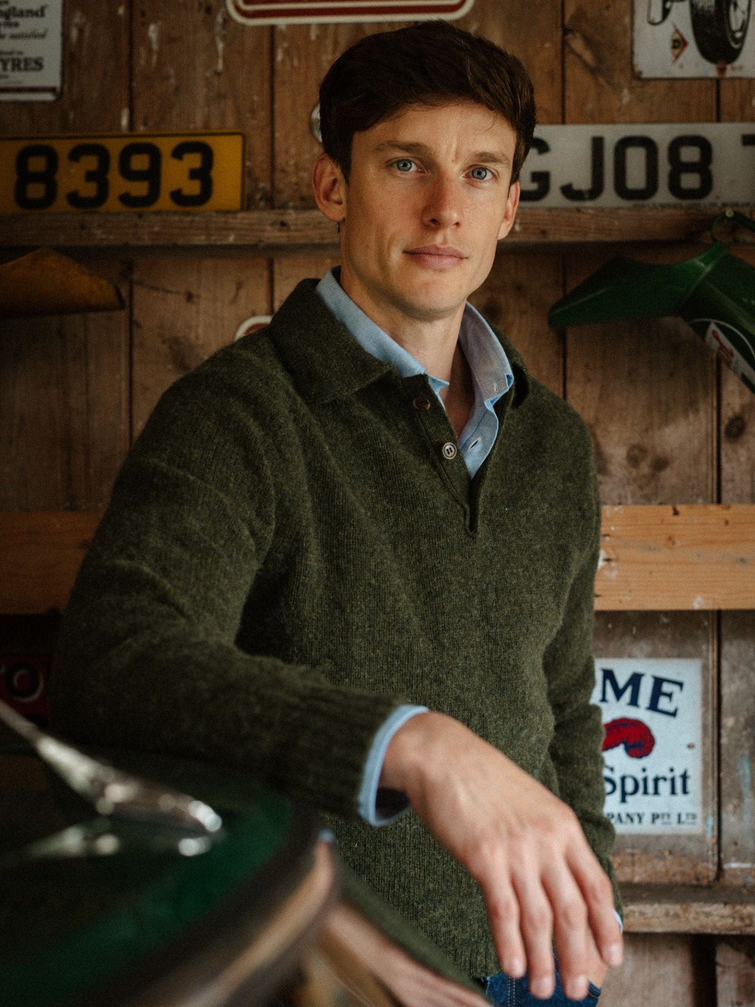 A man with short brown hair, dressed in a Campbell's of Beauly Brushed Shetland Rugby Jumper over a blue collared shirt, stands in a rustic garage adorned with vintage license plates and signs on the wooden wall behind him.