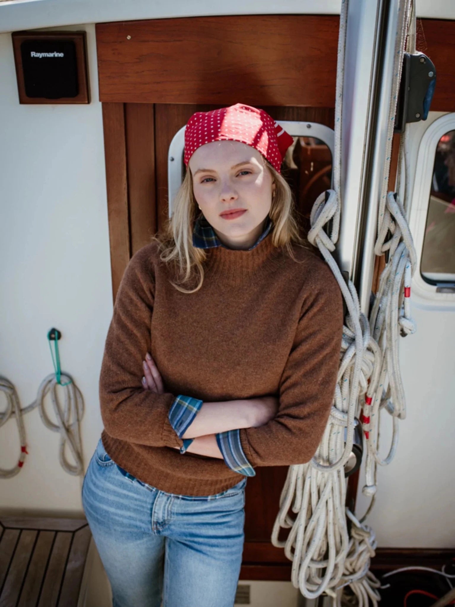 A woman in a red bandana and blue jeans stands on a boat, arms crossed, wearing the Campbell's of Beauly Auld Stock Geelong Lambswool Funnel Neck Jumper, leaning against a wall with coiled ropes beside her.