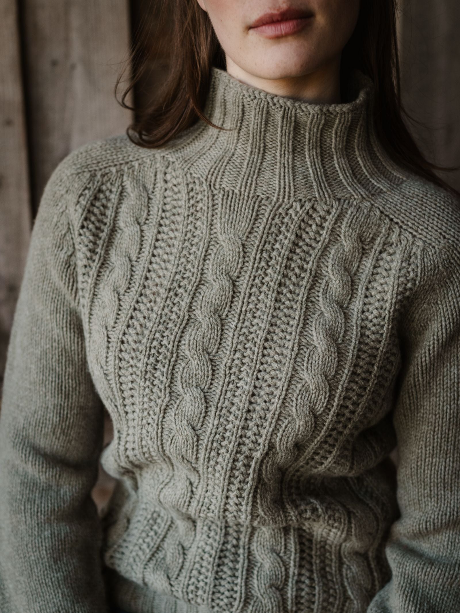 A person models the Campbell's of Beauly Cable Roll Neck Jumper—light gray, supersoft wool, knitted in Scotland with textured cable patterns—shown from shoulders to waist against a wooden background.