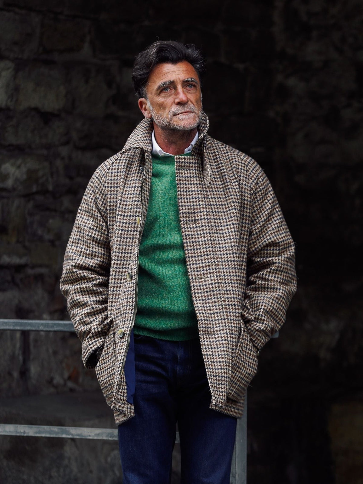 A man with gray hair and beard stands with hands in his pockets in front of a stone wall, wearing the Cannich Coat by Campbell’s of Beauly over a green sweater and jeans, evoking a timeless country style.