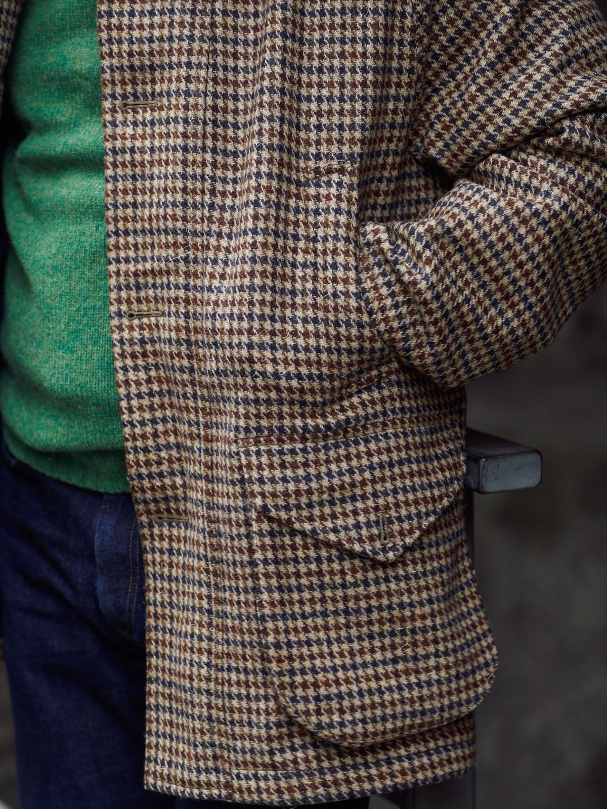A person wearing a green sweater, Campbell’s of Beauly Cannich Coat in brown and beige houndstooth wool, and dark jeans stands with one hand in their coat pocket.