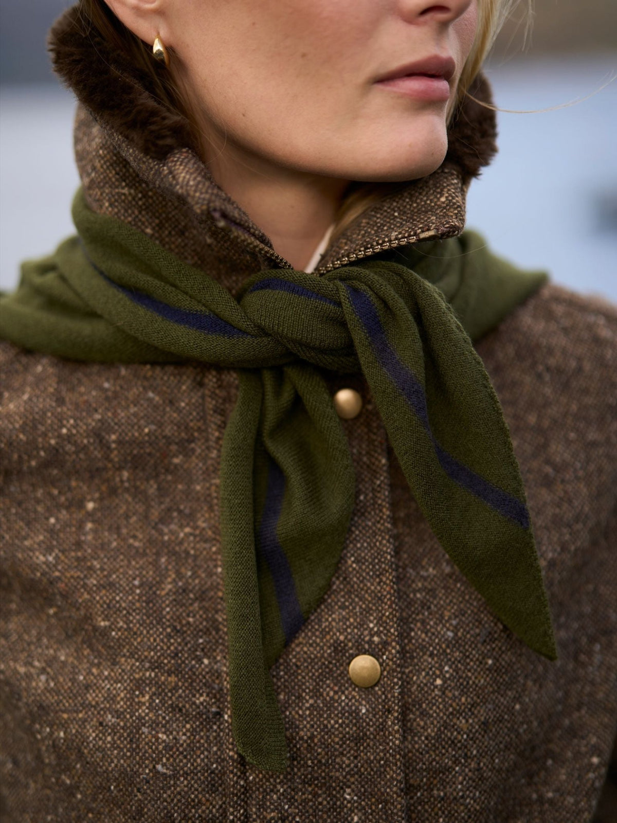 A person wears a dark green, lightweight Campbell’s of Beauly Cashmere Neckerchief with a thin navy blue stripe, tied in a knot over a brown tweed jacket.