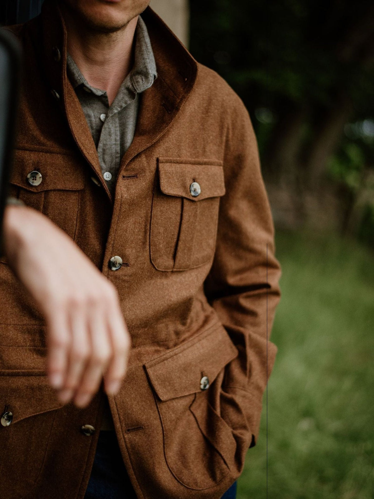 A person wearing the Campbell’s of Beauly Field Teba Jacket in brown and a gray shirt stands outdoors with one arm extended, their face partially out of frame. The blurred background features greenery.