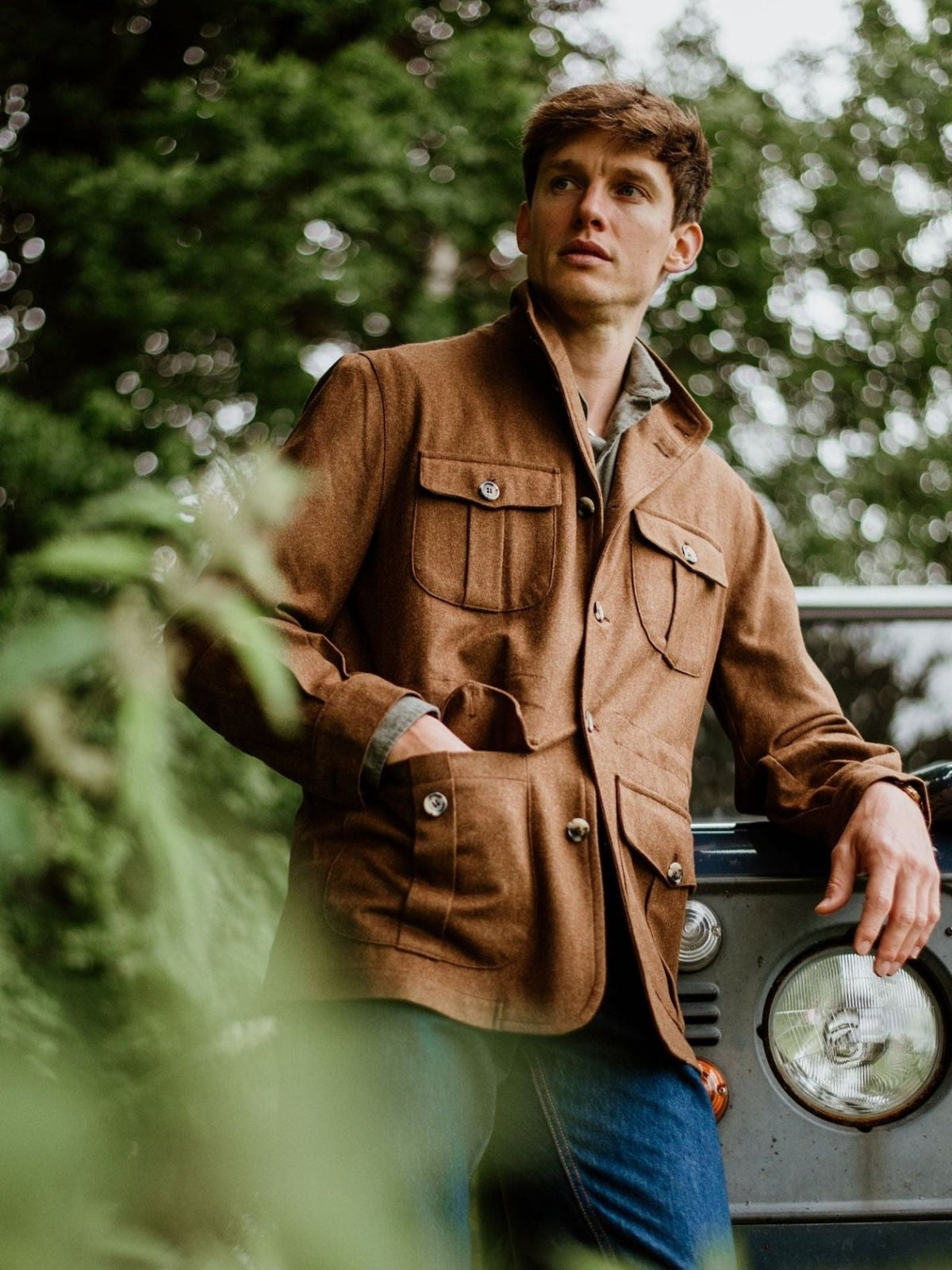 Wearing the Campbell’s of Beauly Field Teba Jacket and blue jeans, a man stands outdoors by a vehicle amidst lush greenery, gazing thoughtfully into the distance.