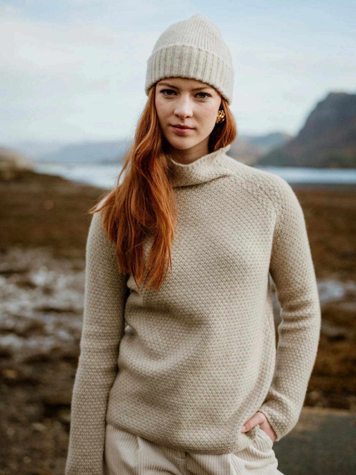 A woman with long red hair, wearing a Scottish knit sweater and a Campbell’s of Beauly Lambswool Hat Cream, stands outdoors by a rocky shore with mountains in the background.