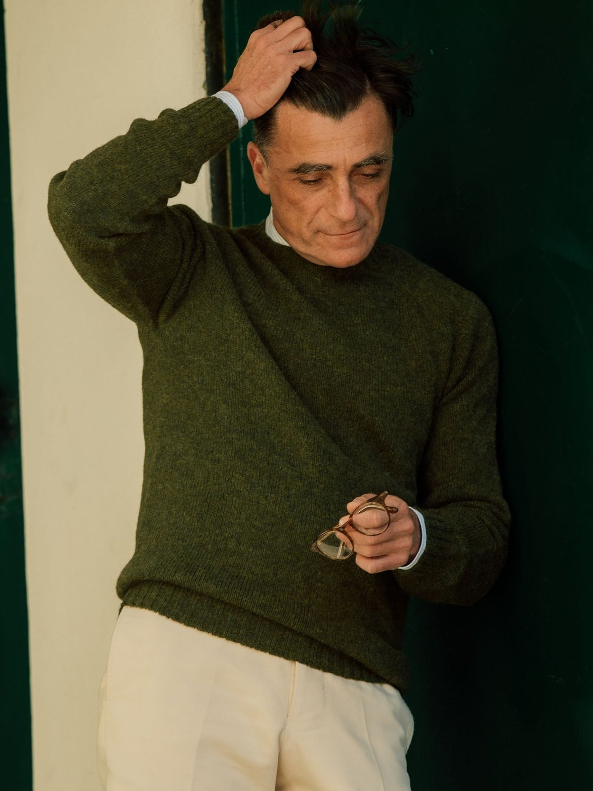 A man wearing the Campbell’s of Beauly Shetland Jumper in green and cream pants stands by a dark green door, holding eyeglasses in one hand and touching his hair with the other, looking down thoughtfully.