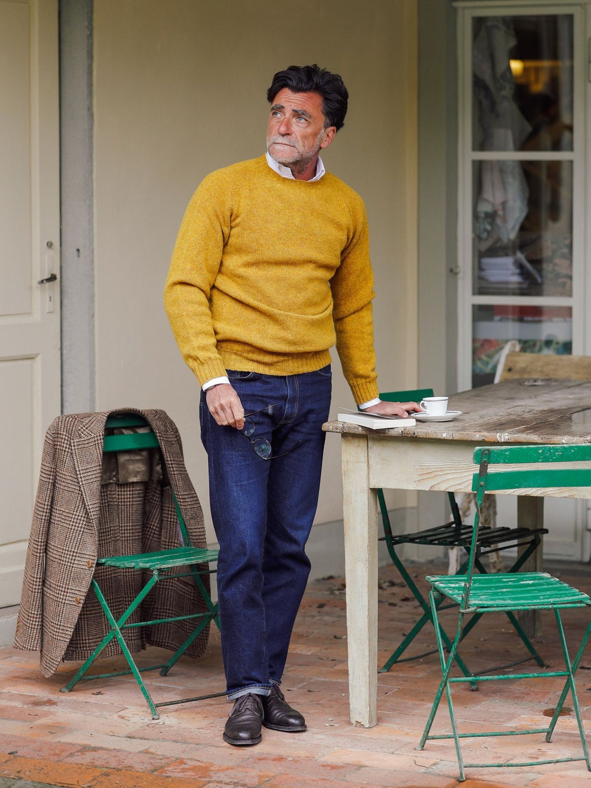 A man in a yellow Shetland Jumper by Campbell’s of Beauly and jeans stands by a rustic wooden table with a cup and saucer. A plaid coat is draped over a green chair, with another green chair nearby. The scene is outdoors on a brick patio.