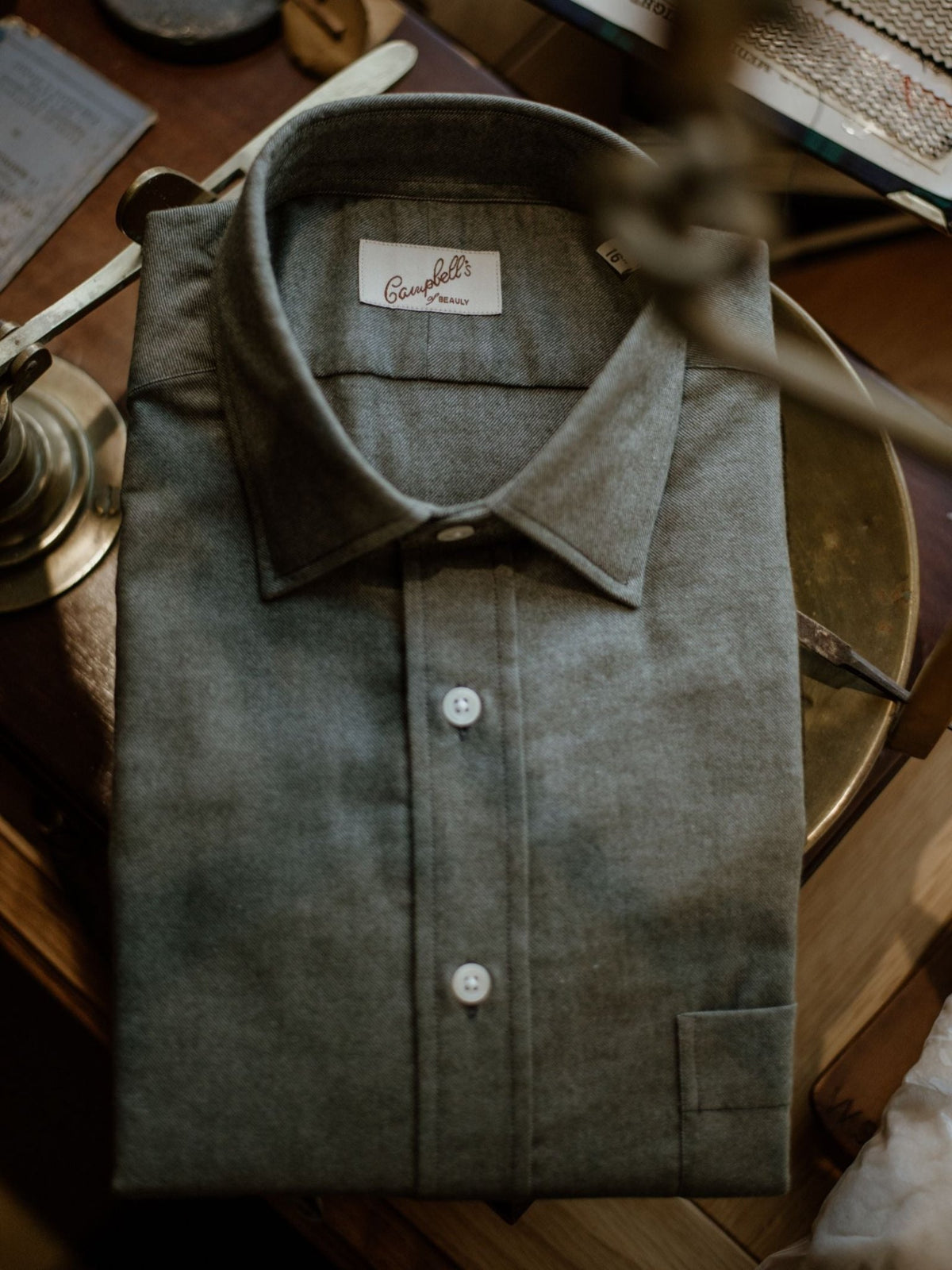 A neatly folded gray Twill Shirt from Campbell’s of Beauly, featuring white buttons, rests on a wooden surface beside a brass lamp.