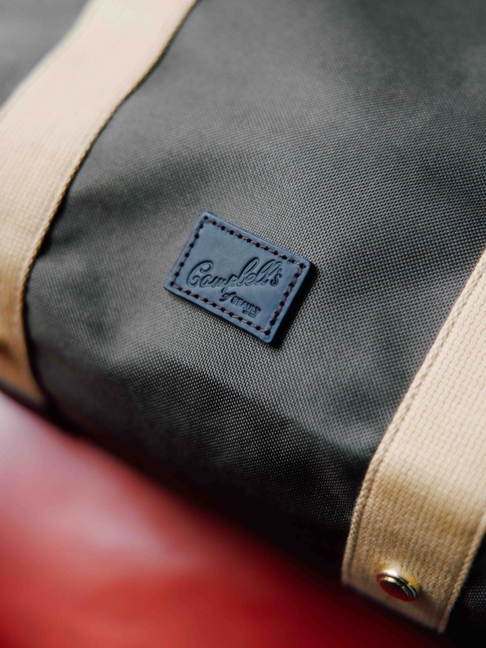 Close-up of the Utility Medium Tote by Campbell's of Beauly in dark green heavy-duty canvas with beige straps, a stitched black Camp Solo patch, and a brass rivet near the strap. The background is blurred red.
