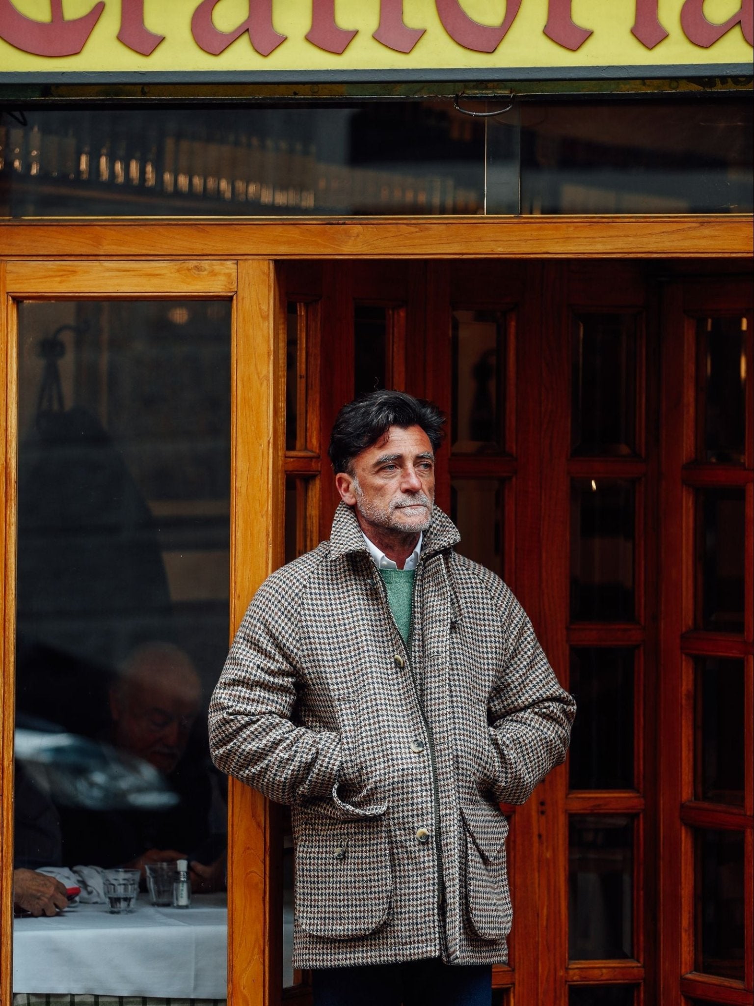 A man with dark hair and a mustache stands outside a wooden door, hands in his pockets, wearing the Cannich Coat by Campbell's of Beauly. Inside the restaurant, someone is seated at a table in the background.
