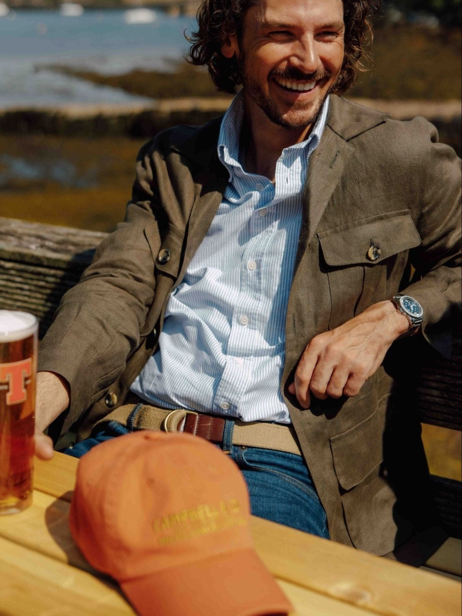 A man sits outdoors smiling, wearing a brown jacket and blue shirt paired with the Arnold Wills & Co Ltd Canvas D-Loop Belt in Natural, holding a pint of beer. An orange cap rests on the wooden table in front of him, perfectly complementing his relaxed country wardrobe.