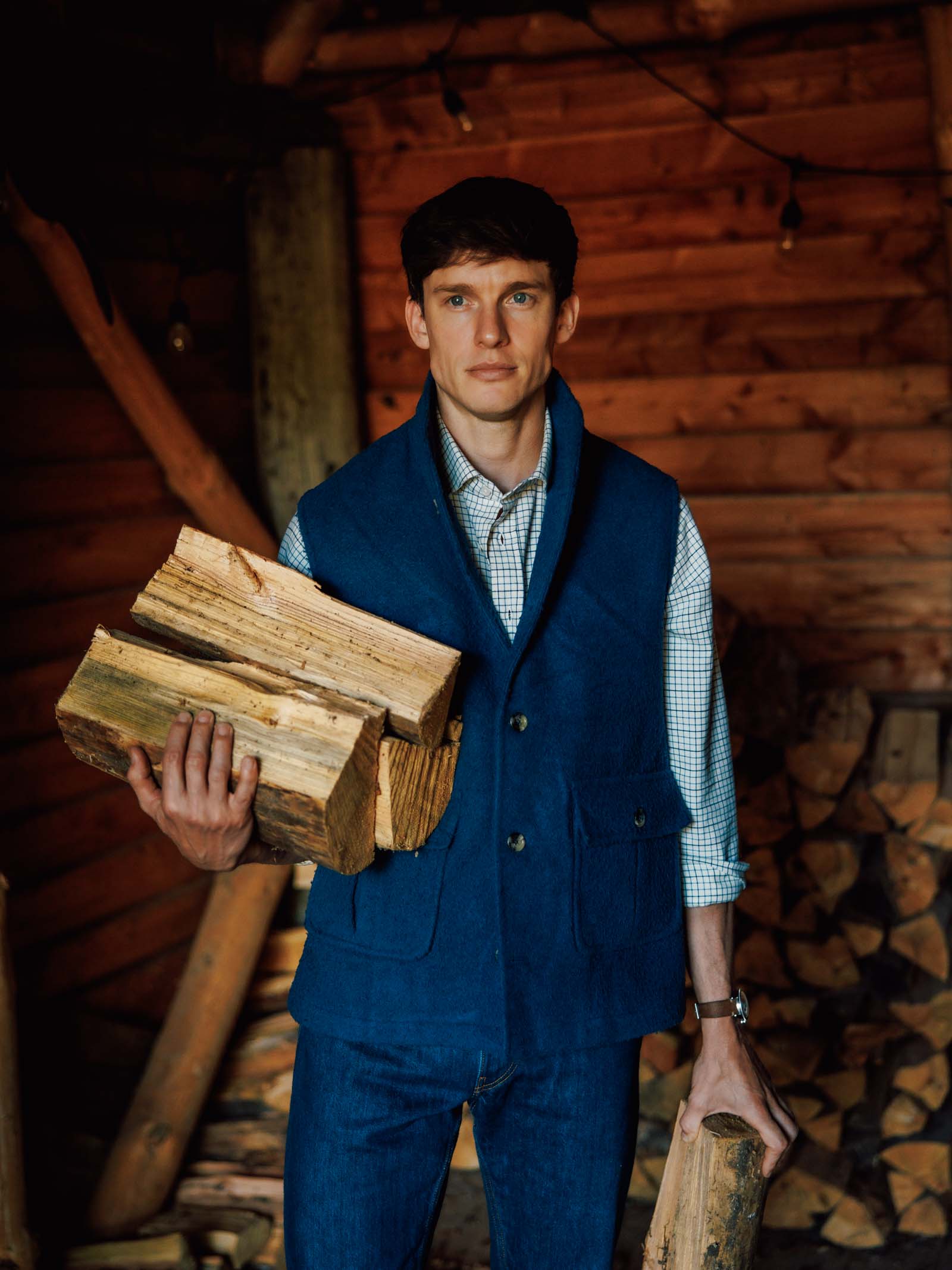 A man in a Campbell’s of Beauly Casentino Fleece Gilet and plaid shirt stands indoors holding firewood, with stacked logs and wooden walls behind him, creating a warm, rustic atmosphere.