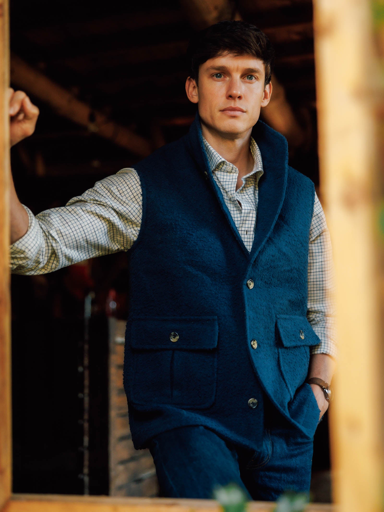 A man stands in a wooden doorway, wearing a blue Campbell's of Beauly Casentino Fleece Gilet over a checked shirt and jeans. He looks ahead with a neutral expression, one arm on the frame as warm light highlights his country style.