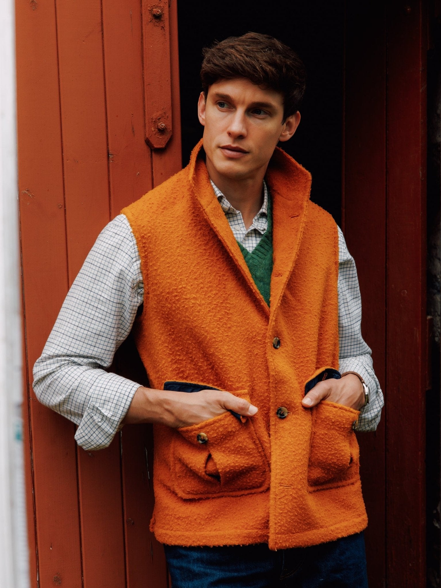 A man stands in a doorway wearing a vibrant orange Campbell's of Beauly Casentino Fleece Gilet over a plaid shirt and green sweater, hands in the gilet’s pockets. He has short brown hair and gazes slightly to the side.