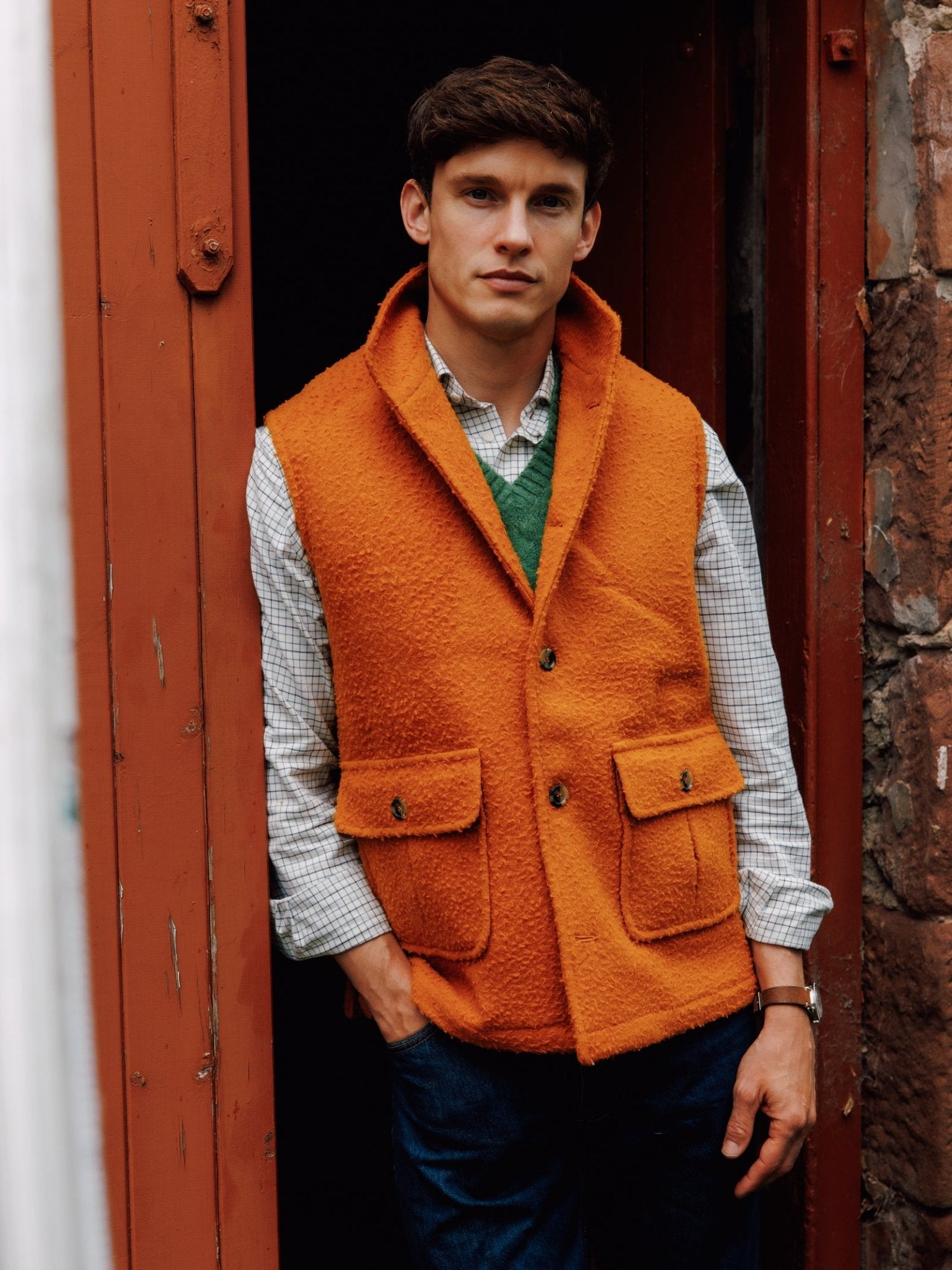 A man with short brown hair stands in a doorway looking at the camera, wearing a bright orange Campbell's of Beauly Casentino Fleece Gilet over a green sweater and checkered shirt, paired with dark blue jeans.