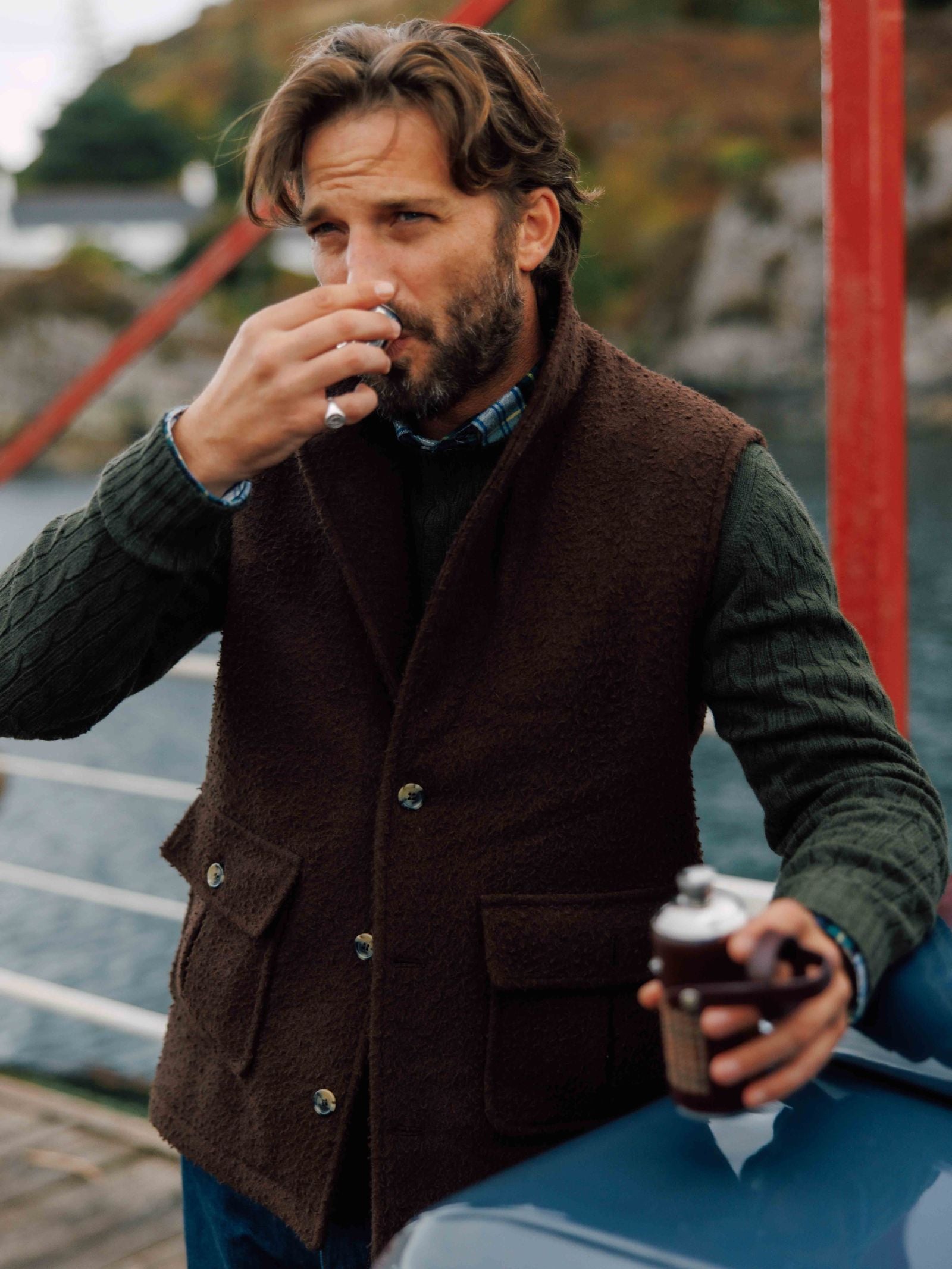 A man with wavy brown hair, wearing a dark brown Casentino Fleece Gilet by Campbell's of Beauly over a green sweater, sips from a small cup and holds a flask outdoors near water and red railings.