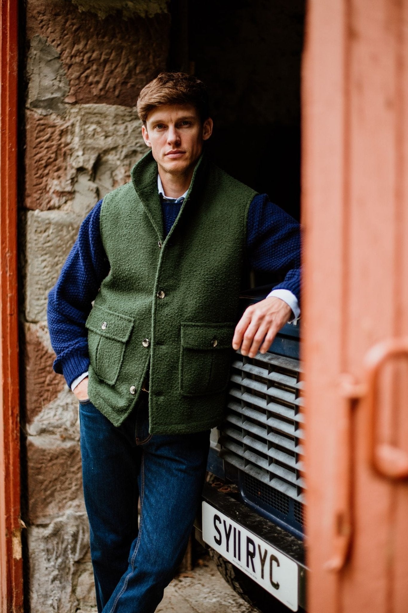 A person in a green Campbell's of Beauly Casentino Fleece Gilet over a blue sweater and jeans leans against a parked vehicle with the license plate SY11 RYC, set against rustic stone walls.