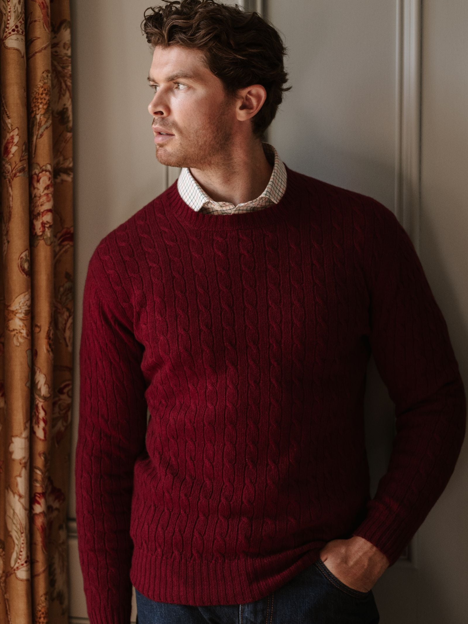 A man with wavy brown hair wears the Campbell's of Beauly Cashmere Cable Crew Jumper in maroon over a collared shirt. He stands indoors by a floral curtain, facing left, with a light wall behind him.