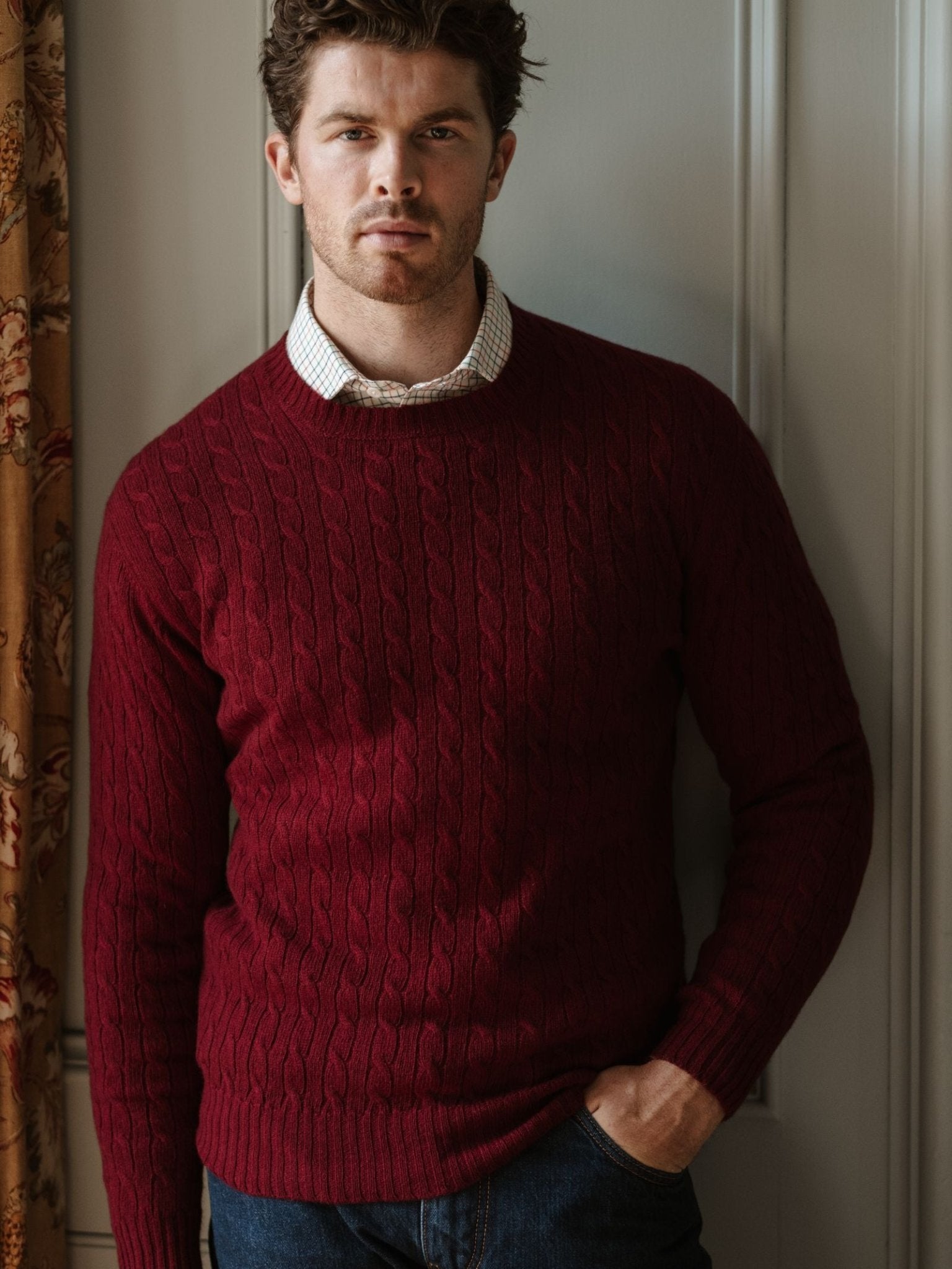 A man with short brown hair and a trimmed beard wears the Campbell's of Beauly Cashmere Cable Crew Jumper in maroon over a white shirt, standing indoors by a floral curtain with one hand in his jeans pocket, looking at the camera.