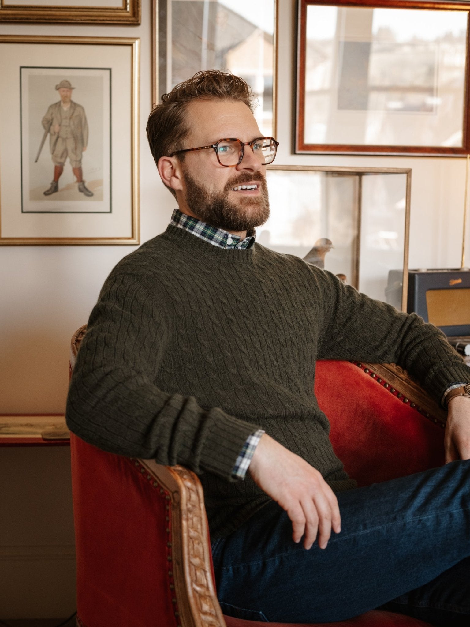 A man with glasses and a beard sits in a red armchair, wearing the Campbell's of Beauly Cashmere Cable Crew Jumper and jeans. Framed art and photos behind him add to the cozy, stylish atmosphere.