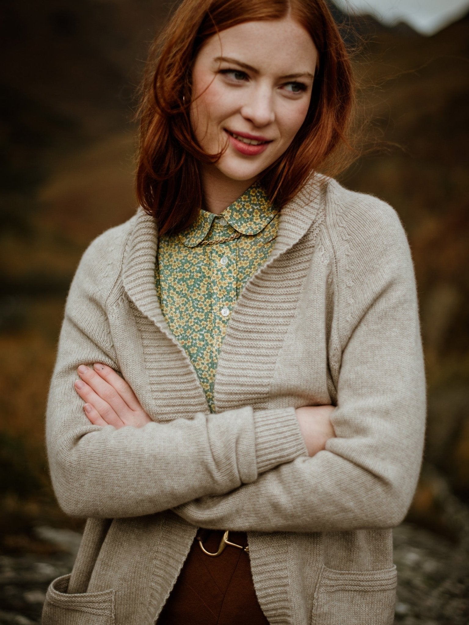 A young woman with long red hair stands outdoors, smiling slightly, wearing the Campbells of Beauly Cashmere Shawl Cardigan over a green floral blouse. Her arms are crossed and the natural background is softly blurred.