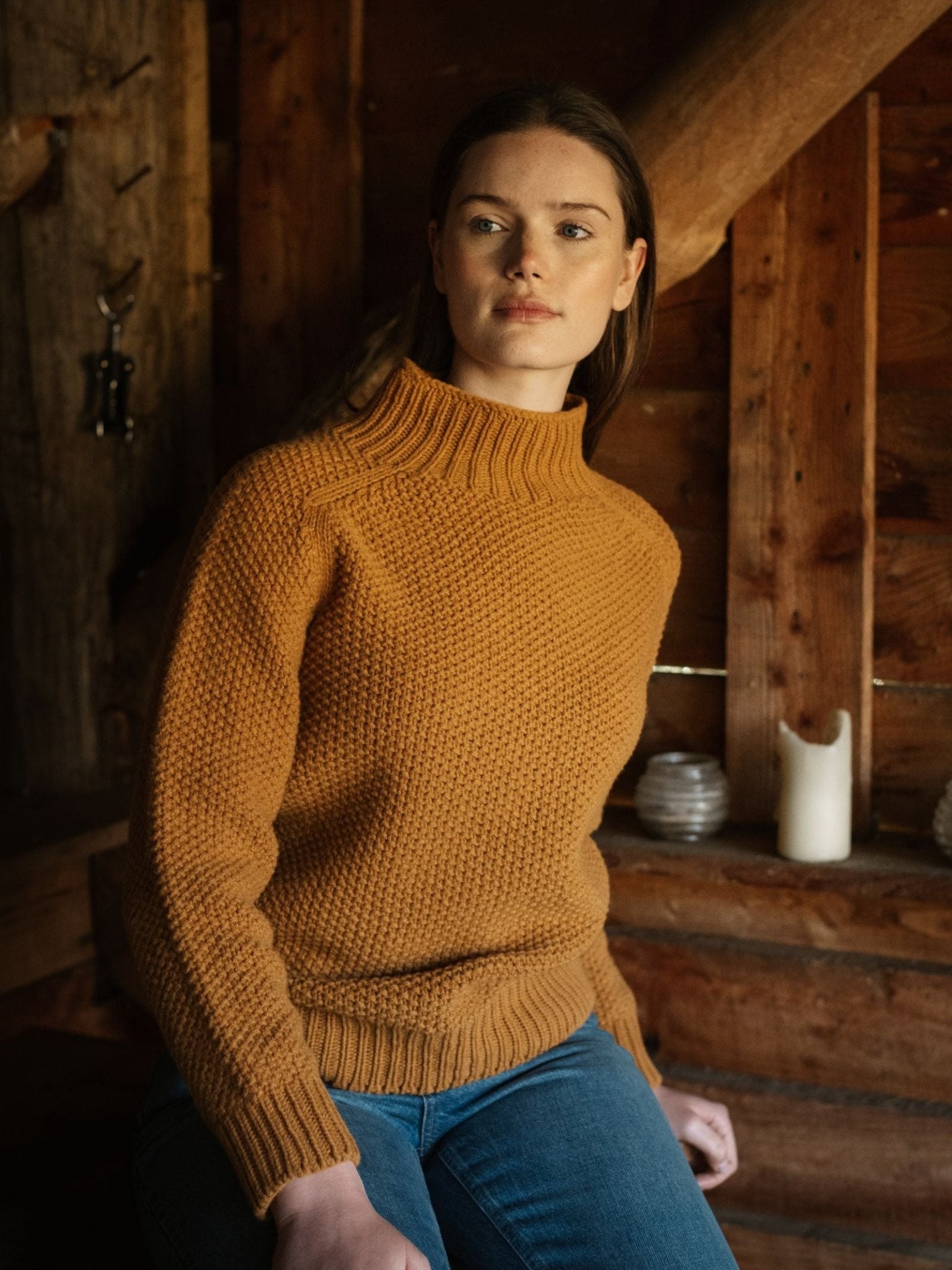 A woman in a Campbell's of Beauly Chunky Moss Stitch Polo and blue jeans sits indoors against a rustic wooden wall with candles and small objects behind her, gazing calmly to the side.