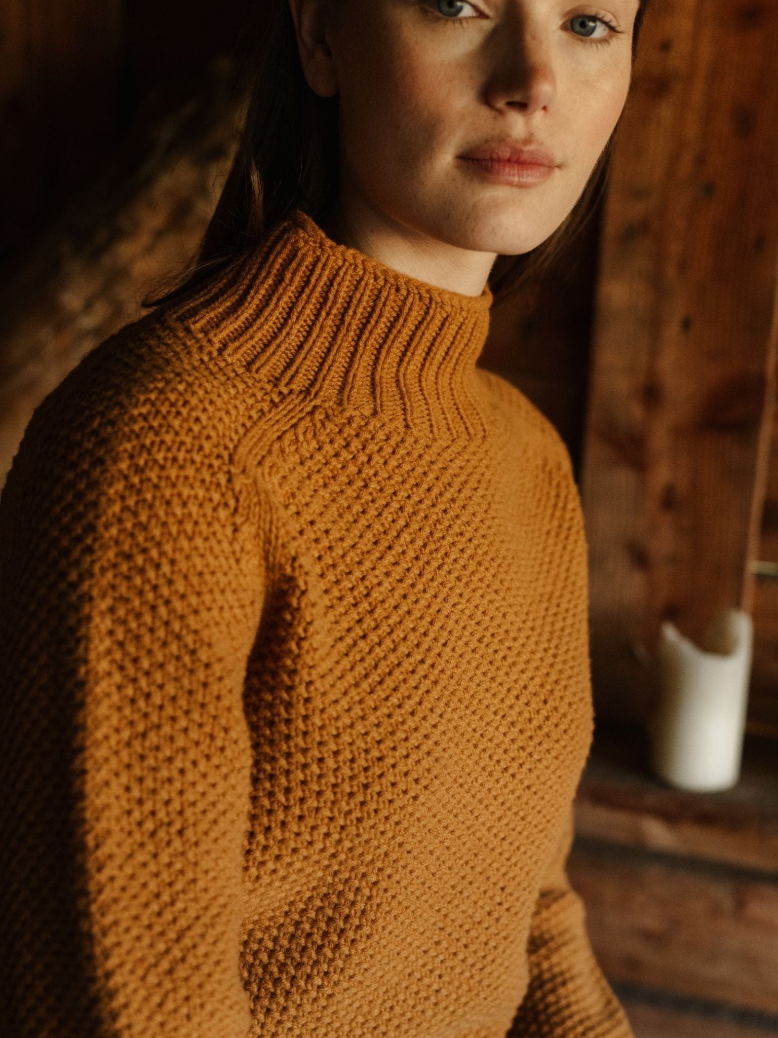 A woman wears the Campbell's of Beauly Chunky Moss Stitch Polo in mustard yellow, sitting indoors by wooden walls and gazing slightly away from the camera with a neutral expression.
