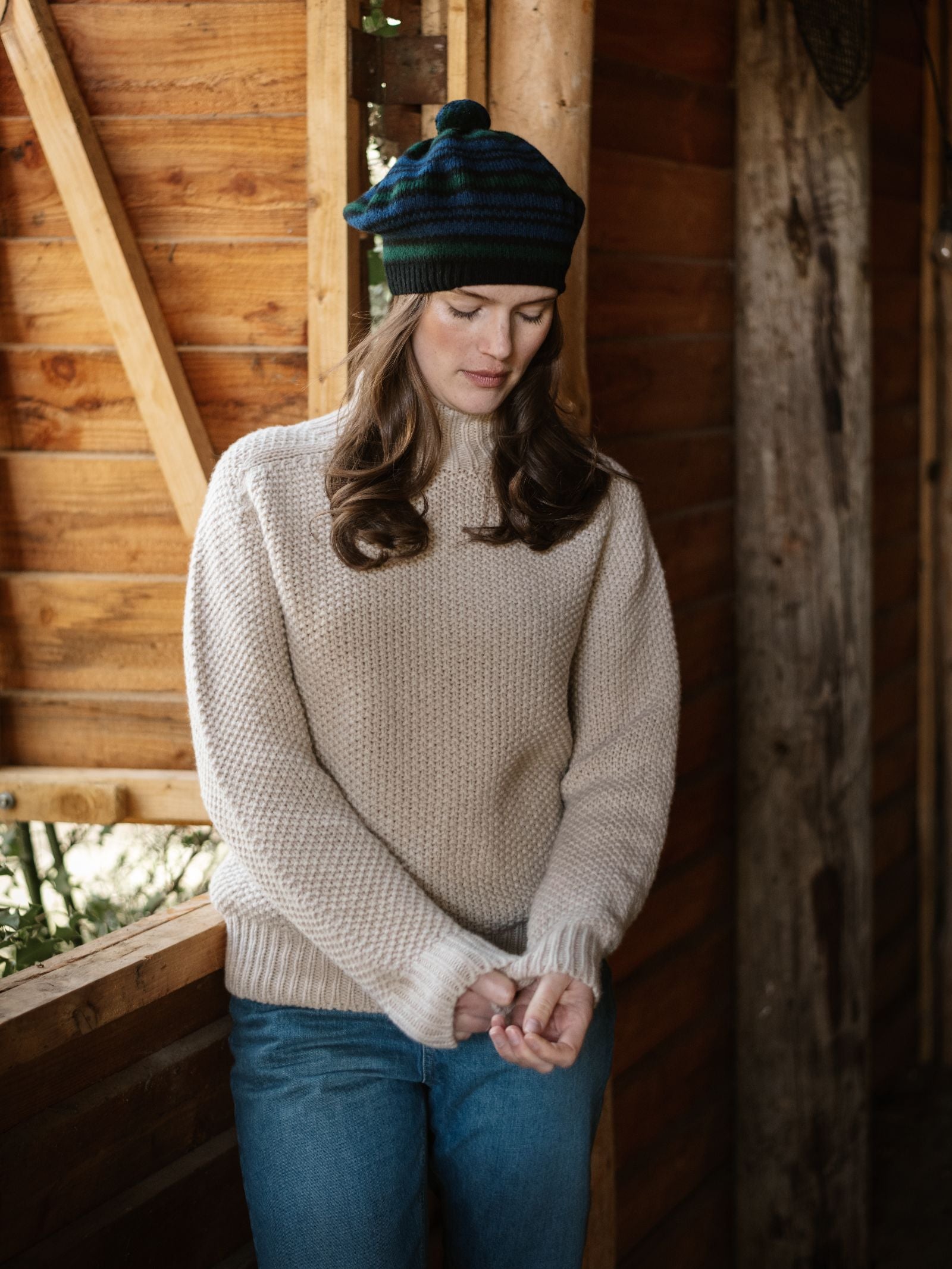 A woman with long brown hair stands indoors by a wooden wall, looking down with hands clasped. She wears the Campbell's of Beauly Chunky Moss Stitch Polo sweater, blue jeans, and a striped knit hat in natural light from a window.