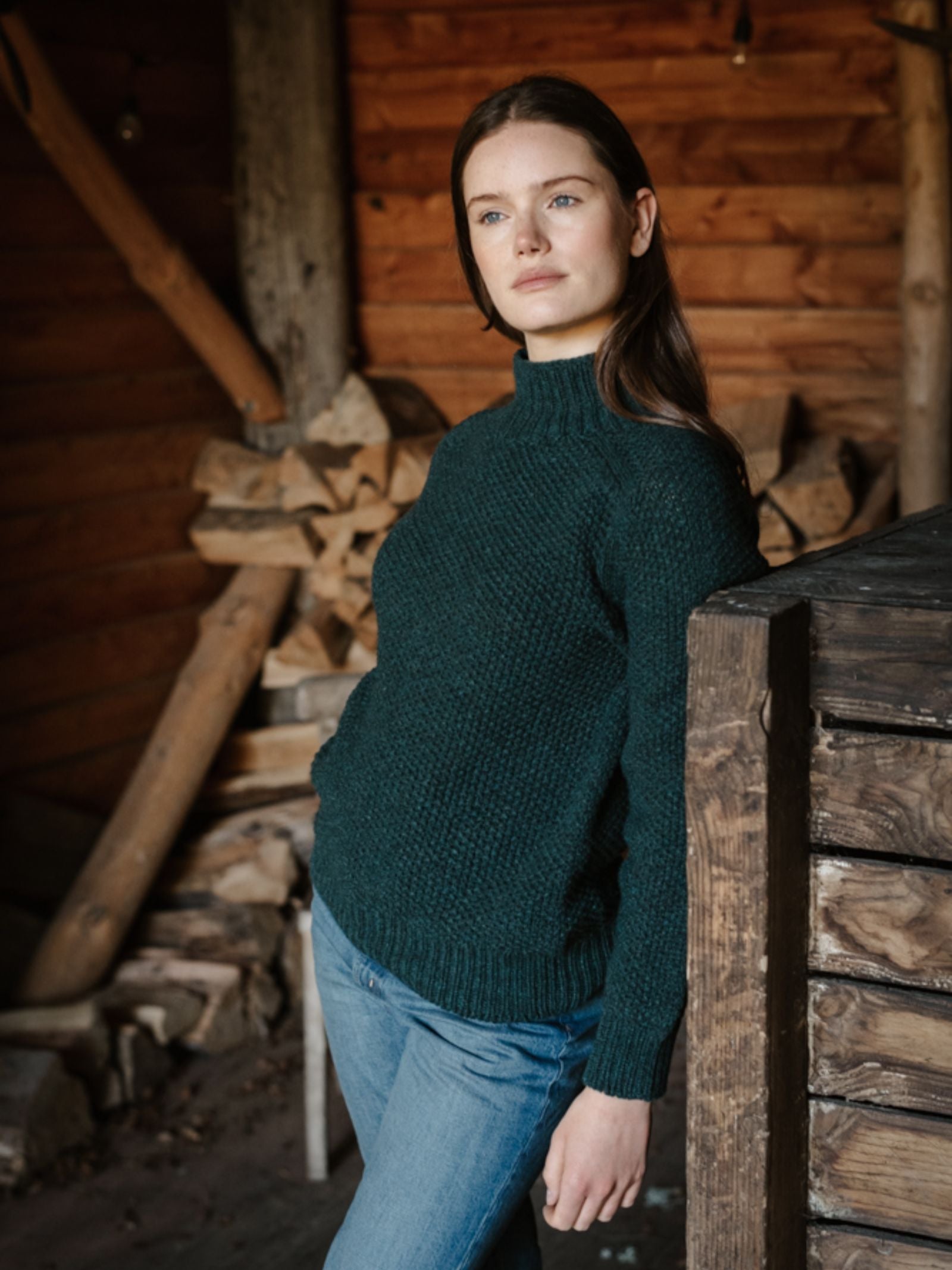A woman with long brown hair, wearing the Campbell's of Beauly Chunky Moss Stitch Polo and blue jeans, leans against a rustic indoor wooden structure with stacked firewood and wooden walls in the background.