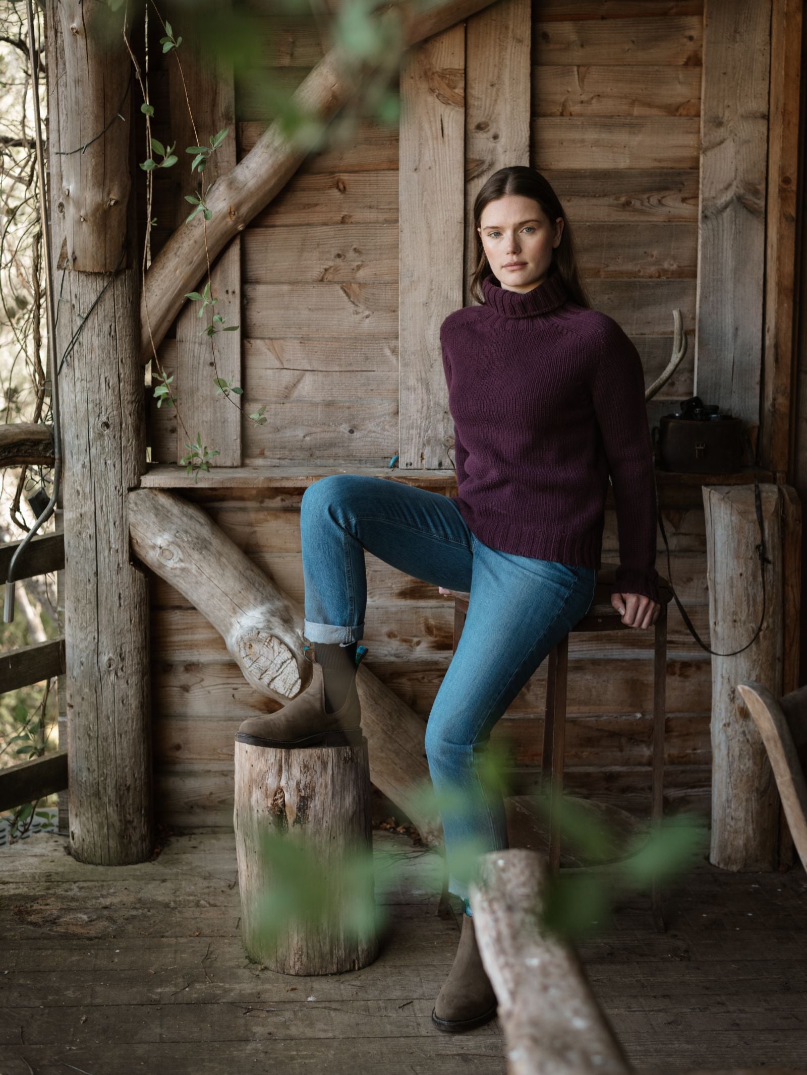 A woman in a Campbell's of Beauly Chunky Roll Neck Sweater in Mulberry and blue jeans poses with one foot on a log inside a rustic wooden cabin, surrounded by wooden walls, beams, and greenery in the foreground.