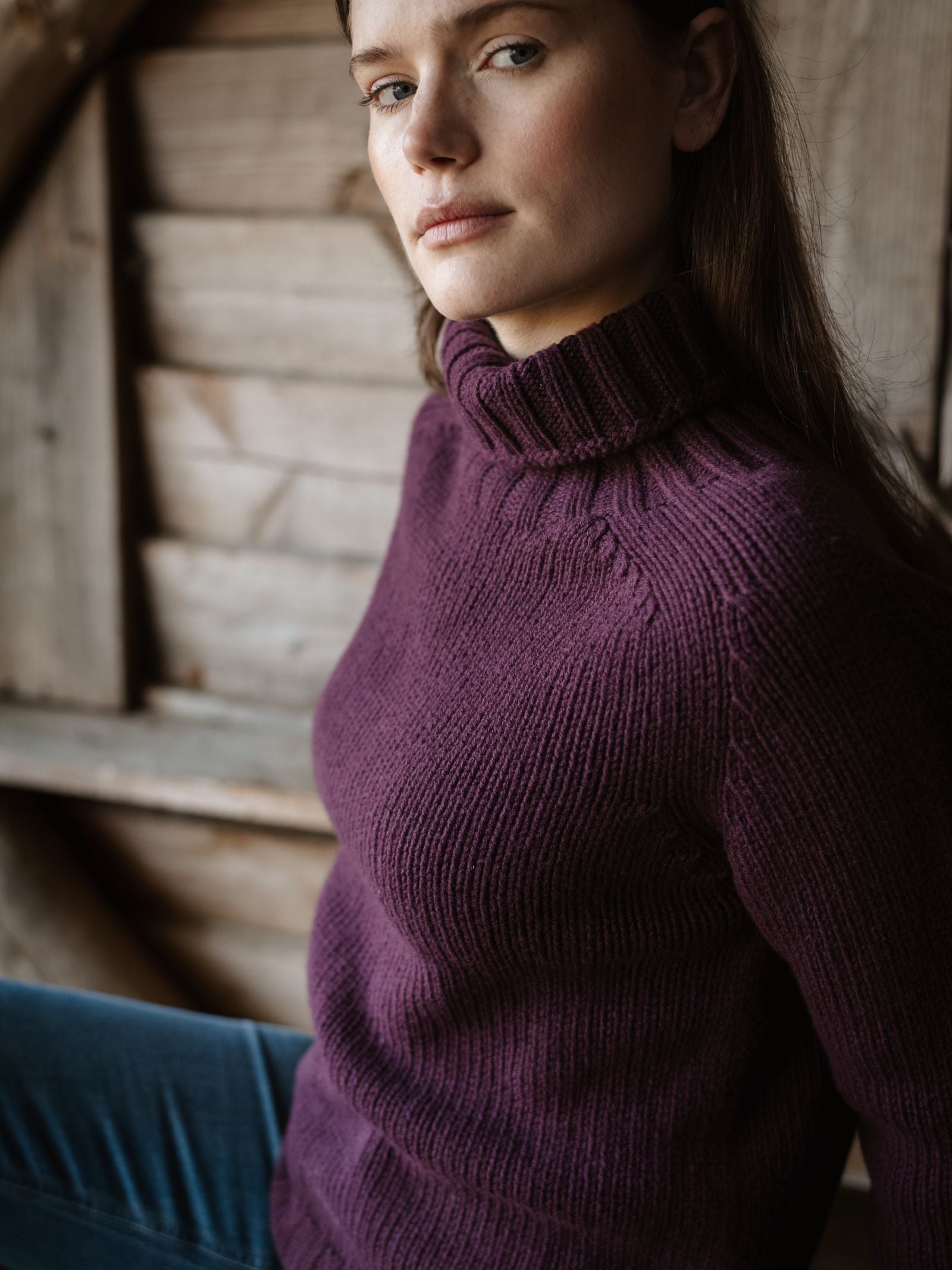 A woman with long brown hair wears the Campbell's of Beauly Chunky Roll Neck Sweater in purple—knitted in Scotland from supersoft wool—sitting indoors by a wooden wall, looking to the side with a calm expression.