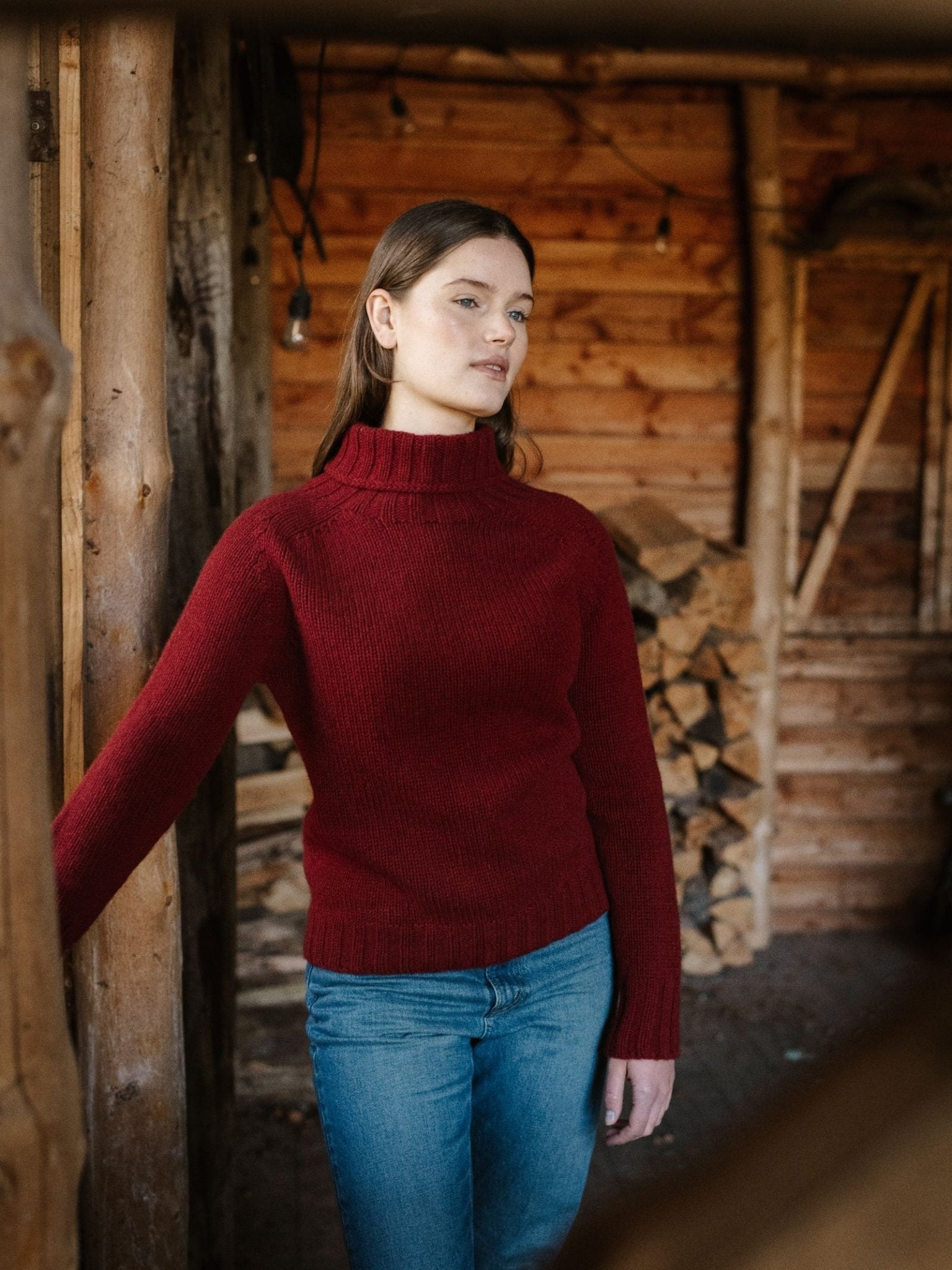 Wearing the Campbell's of Beauly Chunky Roll Neck Sweater in ruby and blue jeans, a woman stands indoors near a wooden wall and stacked firewood, gazing calmly away in a cozy, rustic setting.