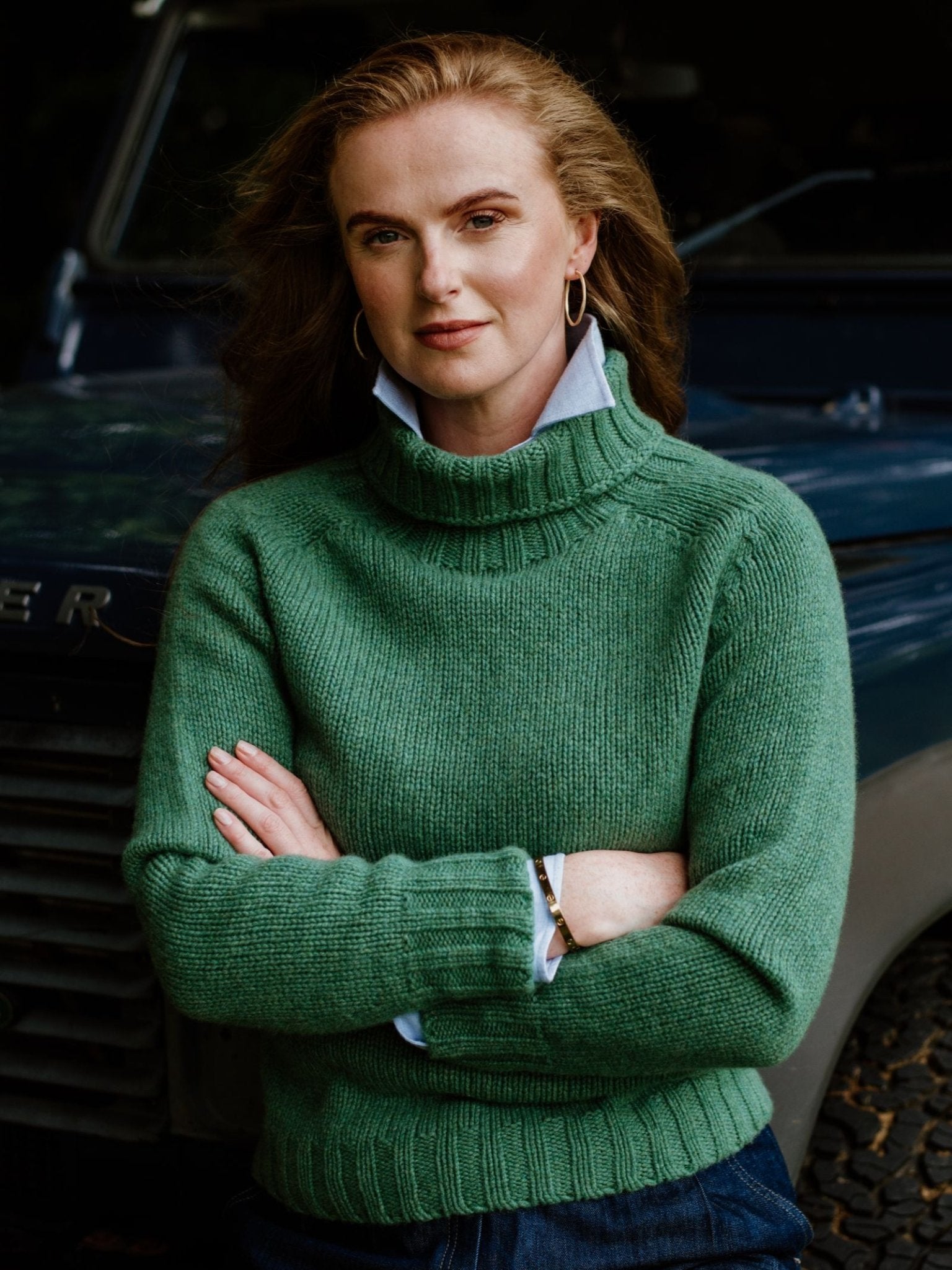 A woman with long, wavy hair stands confidently with crossed arms in front of a dark vehicle, modeling the Campbell's of Beauly Chunky Roll Neck Sweater.