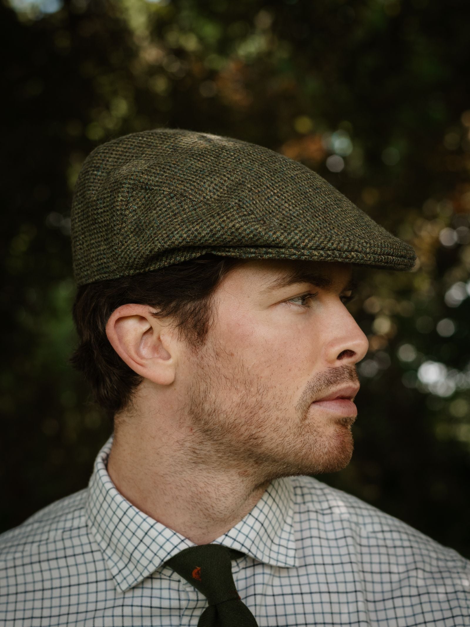 A man with light skin and short brown hair wears the Unisex Green Conon Cap by Campbell's of Beauly, a white checkered shirt, and a green tie. He is outdoors in side profile with a blurred, leafy background.