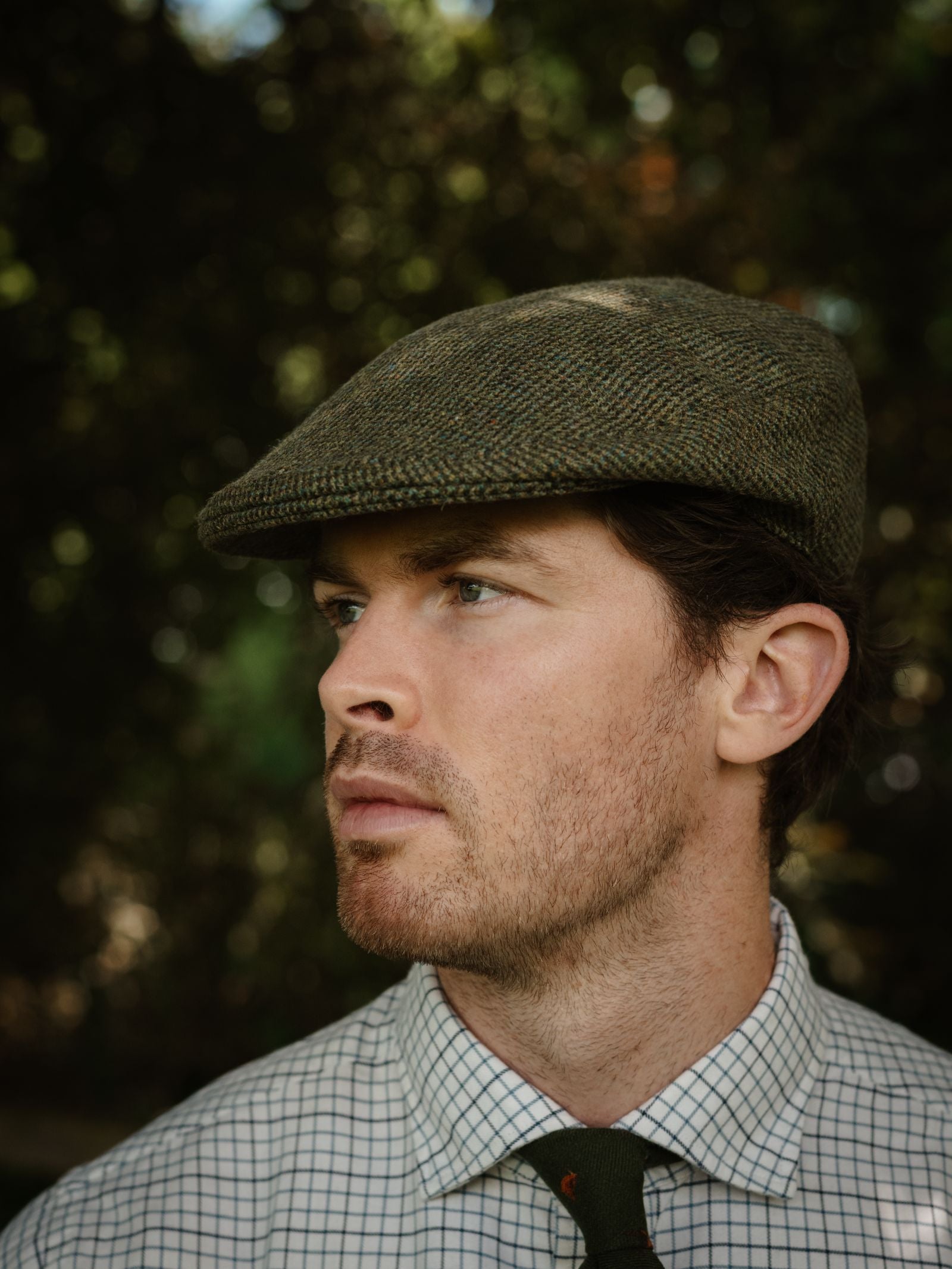 A person wearing the Campbell's of Beauly Unisex Green Conon Cap, a checkered shirt, and a dark green tie looks to the side against a softly blurred outdoor backdrop with trees and dappled sunlight.