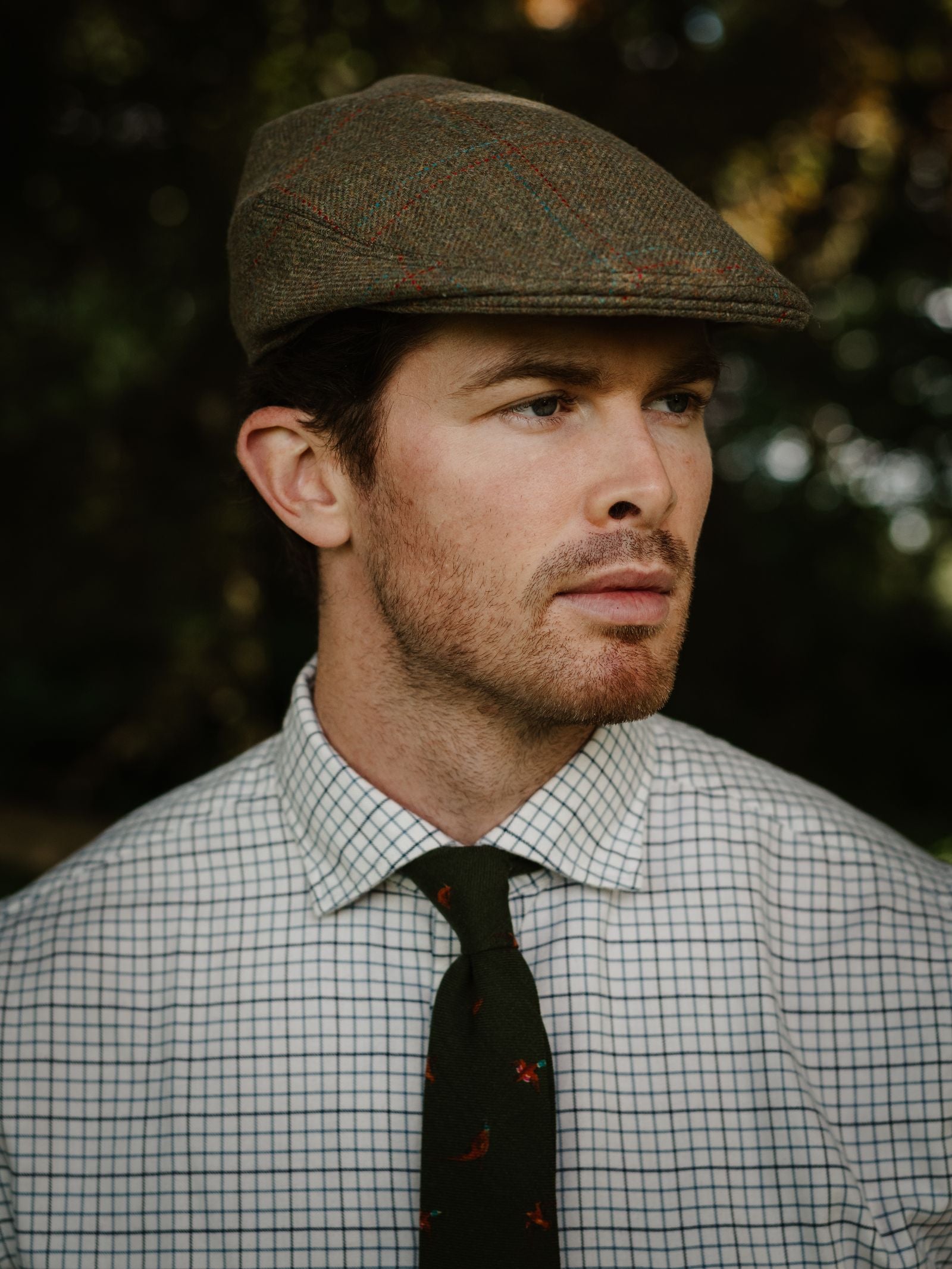 A man with short brown hair and a trimmed beard wears the Unisex Brown Conon Cap by Campbell's of Beauly, paired with a white checkered shirt and dark patterned tie, standing outdoors against a blurred natural background.