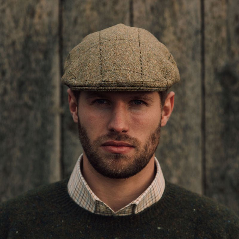 A man with a short beard wears the Auld Stock Conon Cap from Campbell's of Beauly, paired with a green sweater and plaid collared shirt, standing before a rustic wooden backdrop.