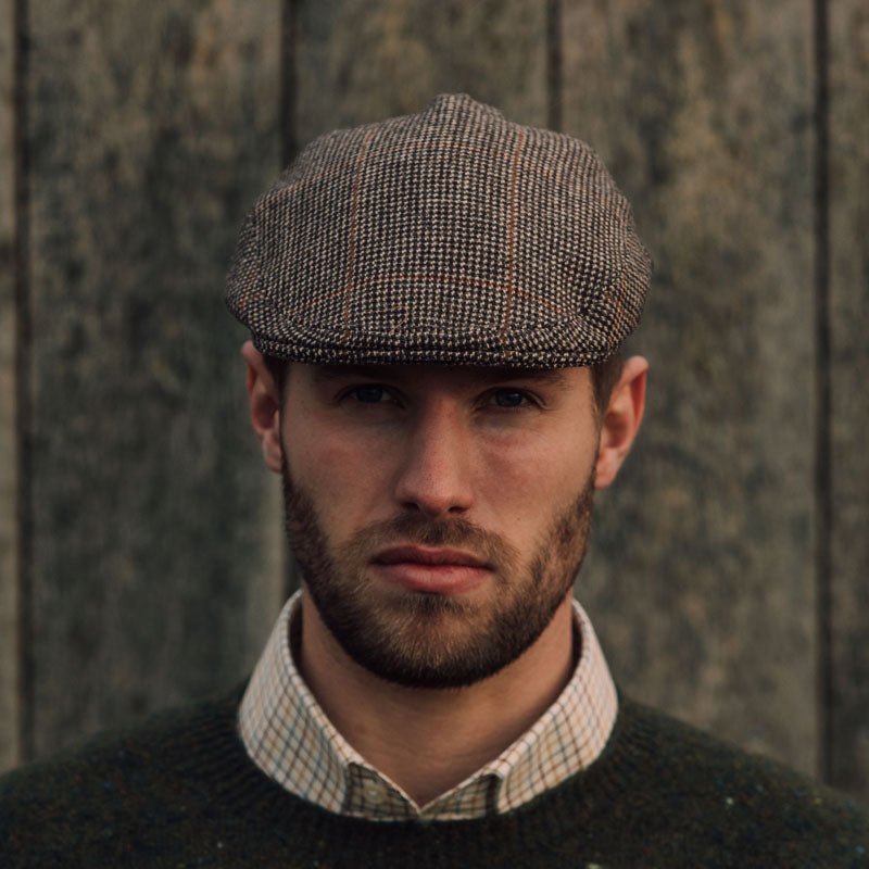A bearded man wears the Campbell's of Beauly Unisex Brown Conon Cap, paired with a green sweater and checked shirt, standing before a weathered wooden background.