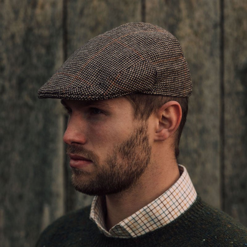 A man with a trimmed beard wears the Campbell's of Beauly Unisex Brown Conon Cap in color K523, paired with a green sweater and checkered shirt, as he stands before a weathered wooden wall.