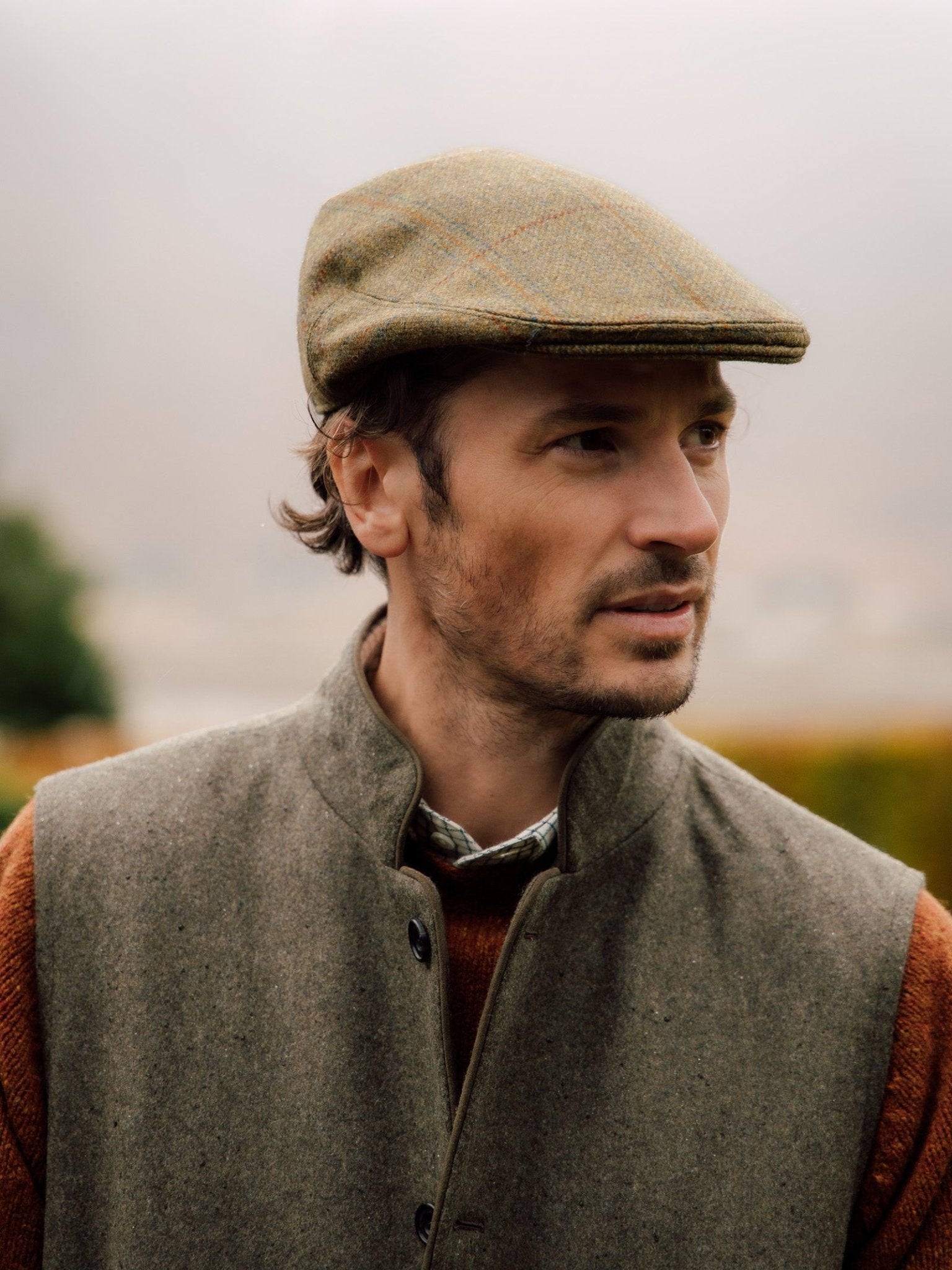 A man in a Campbell's of Beauly Auld Stock Conon Cap stands outdoors in soft, natural light and earthy tones, the blurred backdrop highlighting the timeless British tweed heritage of his flat cap.