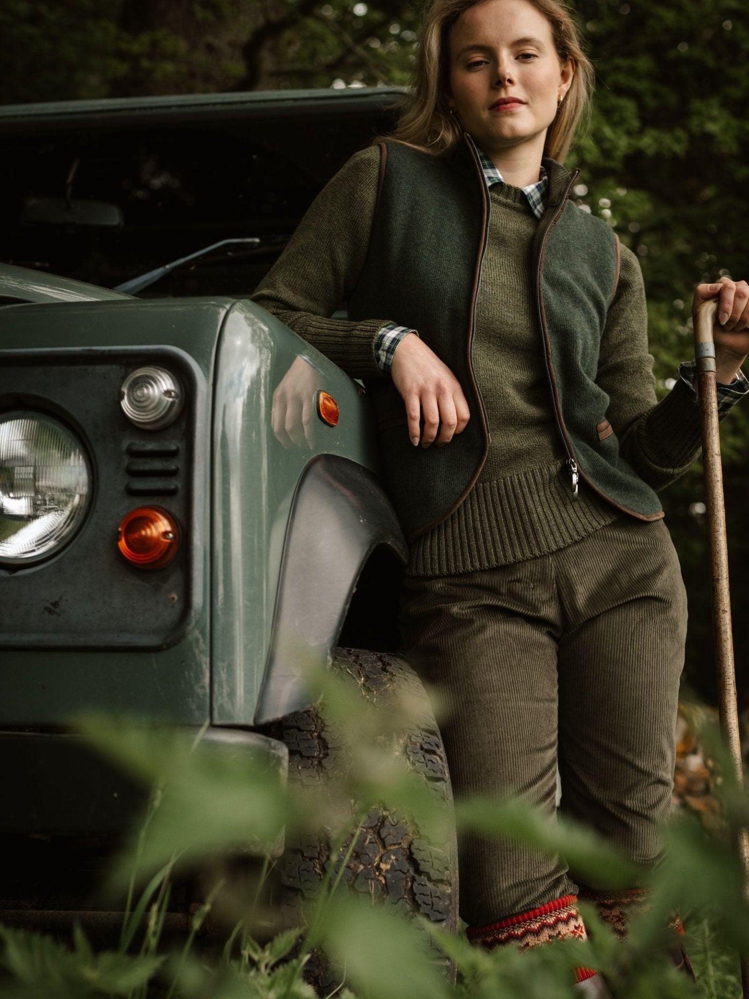 A woman wearing Campbell's of Beauly Corduroy Plus Twos leans on a green SUV with a walking stick, surrounded by woodland foliage and trees.