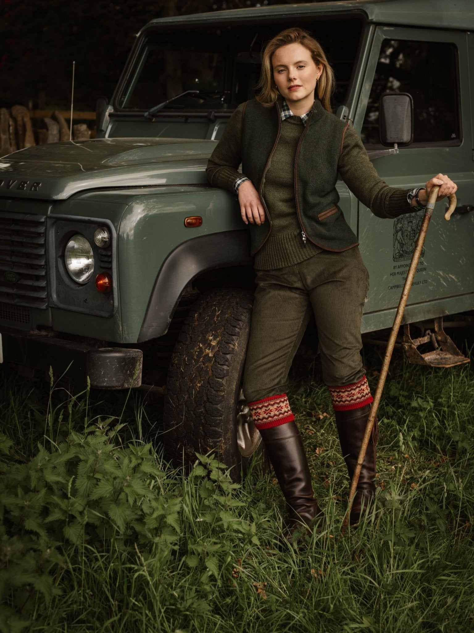 A woman wearing Corduroy Plus Twos by Campbell's of Beauly leans on a walking stick beside a green Land Rover parked on grass, with bushes and trees in the background.