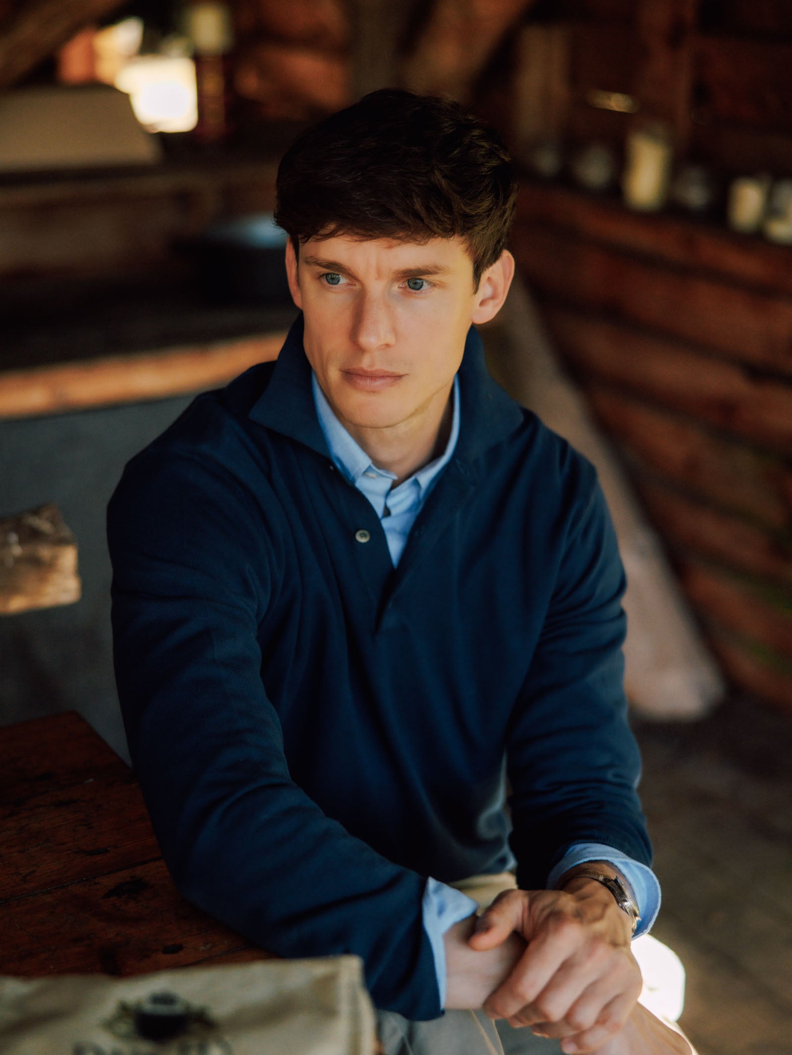 A young man with short brown hair sits indoors at a wooden table, thoughtfully gazing to the side, wearing a blue Cotton Rugby from Campbell's of Beauly. The softly lit background features wooden walls and shelves.