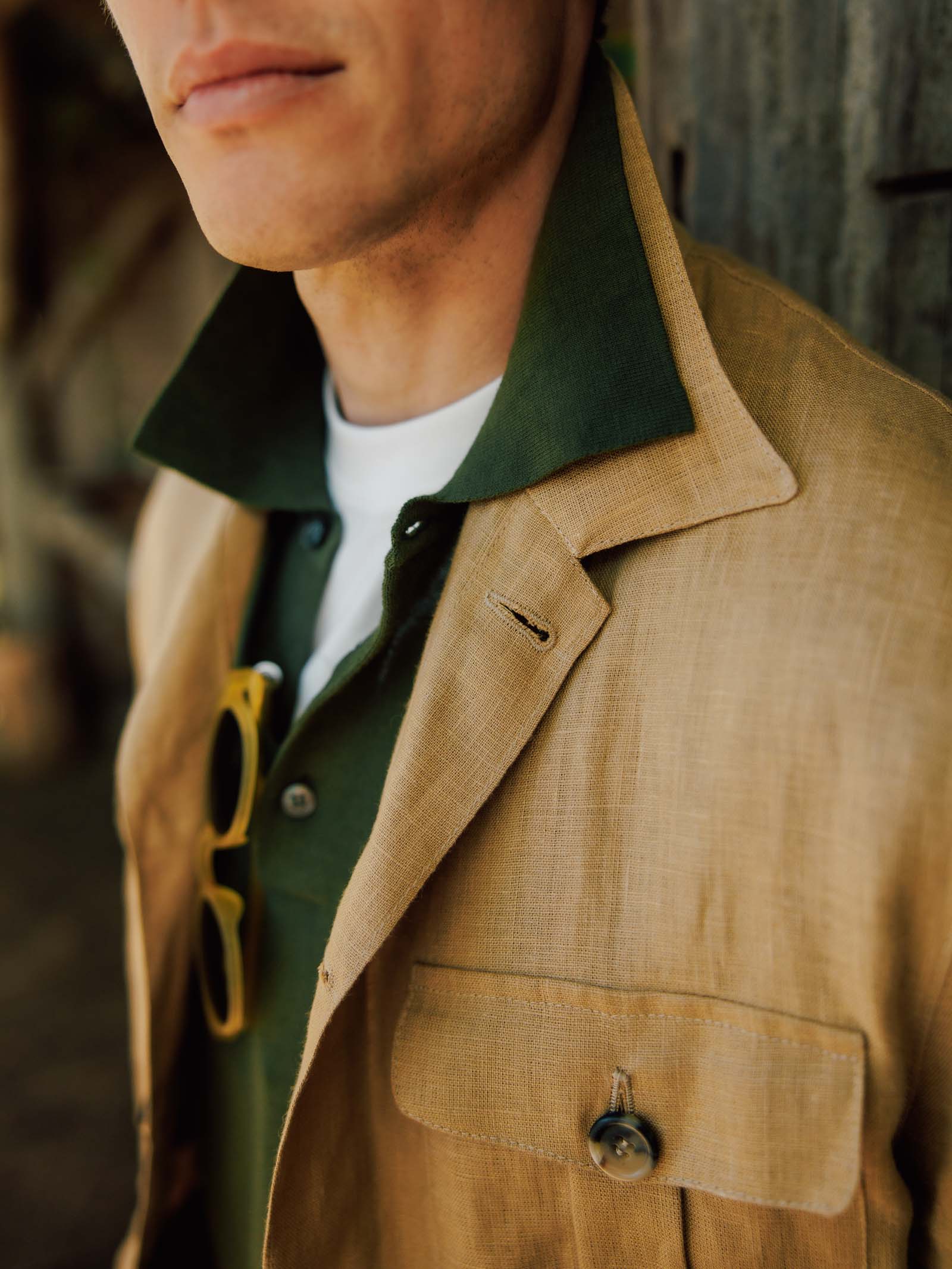 A man wears a Campbell's of Beauly Cotton Rugby in green under a tan linen jacket, with yellow sunglasses hanging from the jacket pocket; the background is blurred.