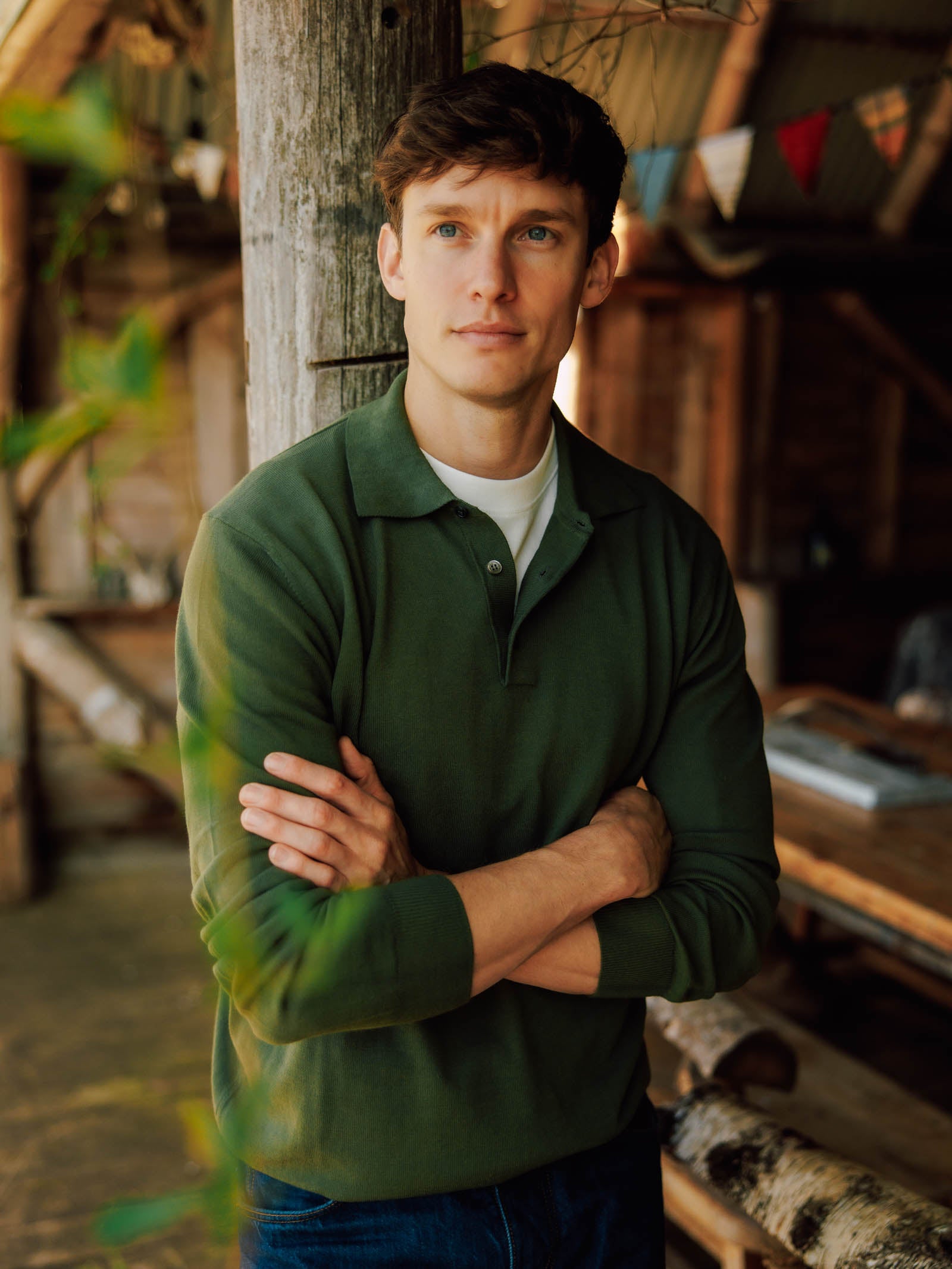 A man with short brown hair wears a green Campbell's of Beauly Cotton Rugby shirt, standing indoors with arms crossed against a wooden post. The rustic scene includes wooden furniture and colorful bunting in the background.