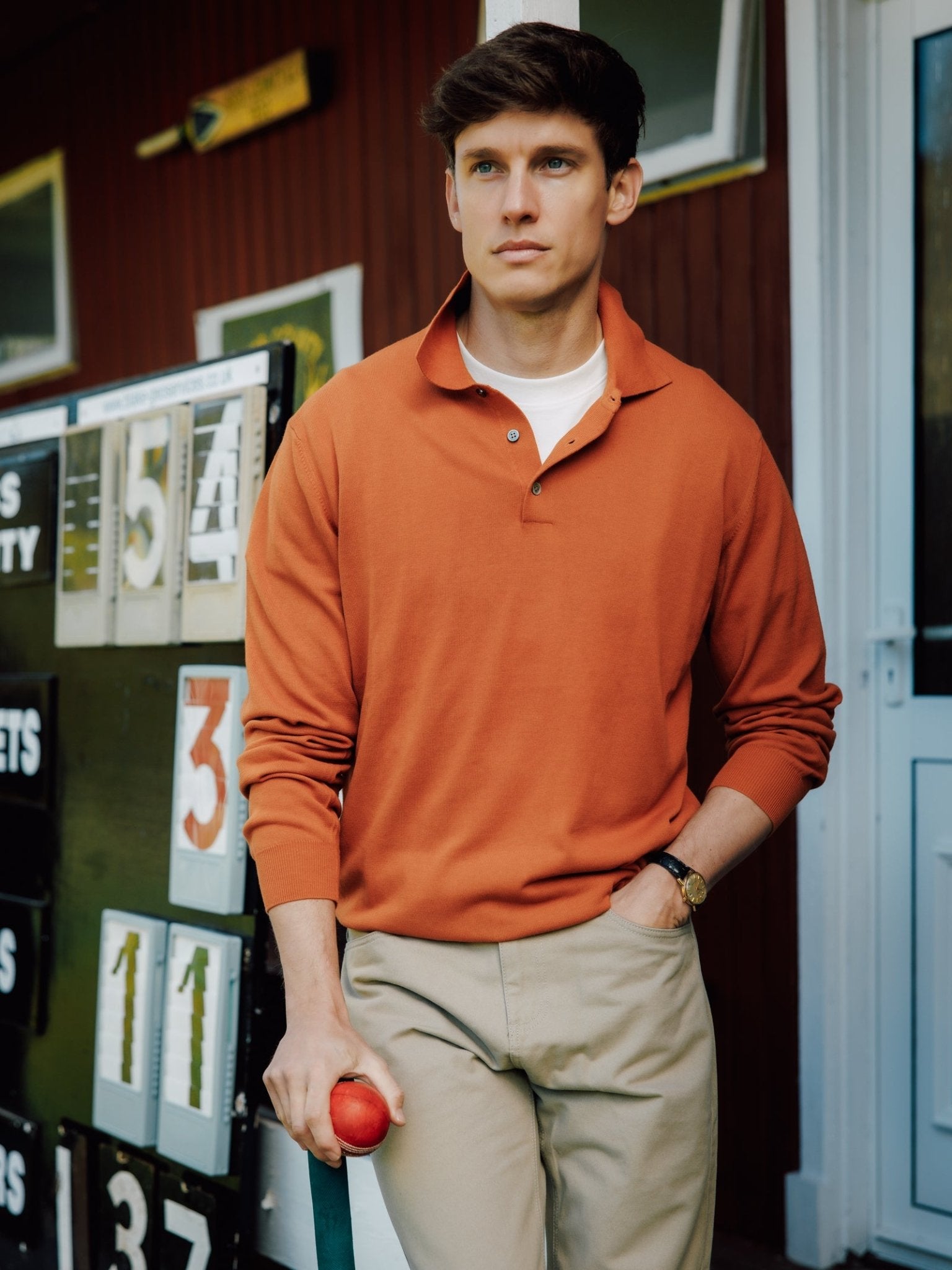 A brown-haired man wears the Campbell's of Beauly Cotton Rugby with beige pants, holding a red ball. Behind him are a scoreboard showing numbers and a red building with windows.