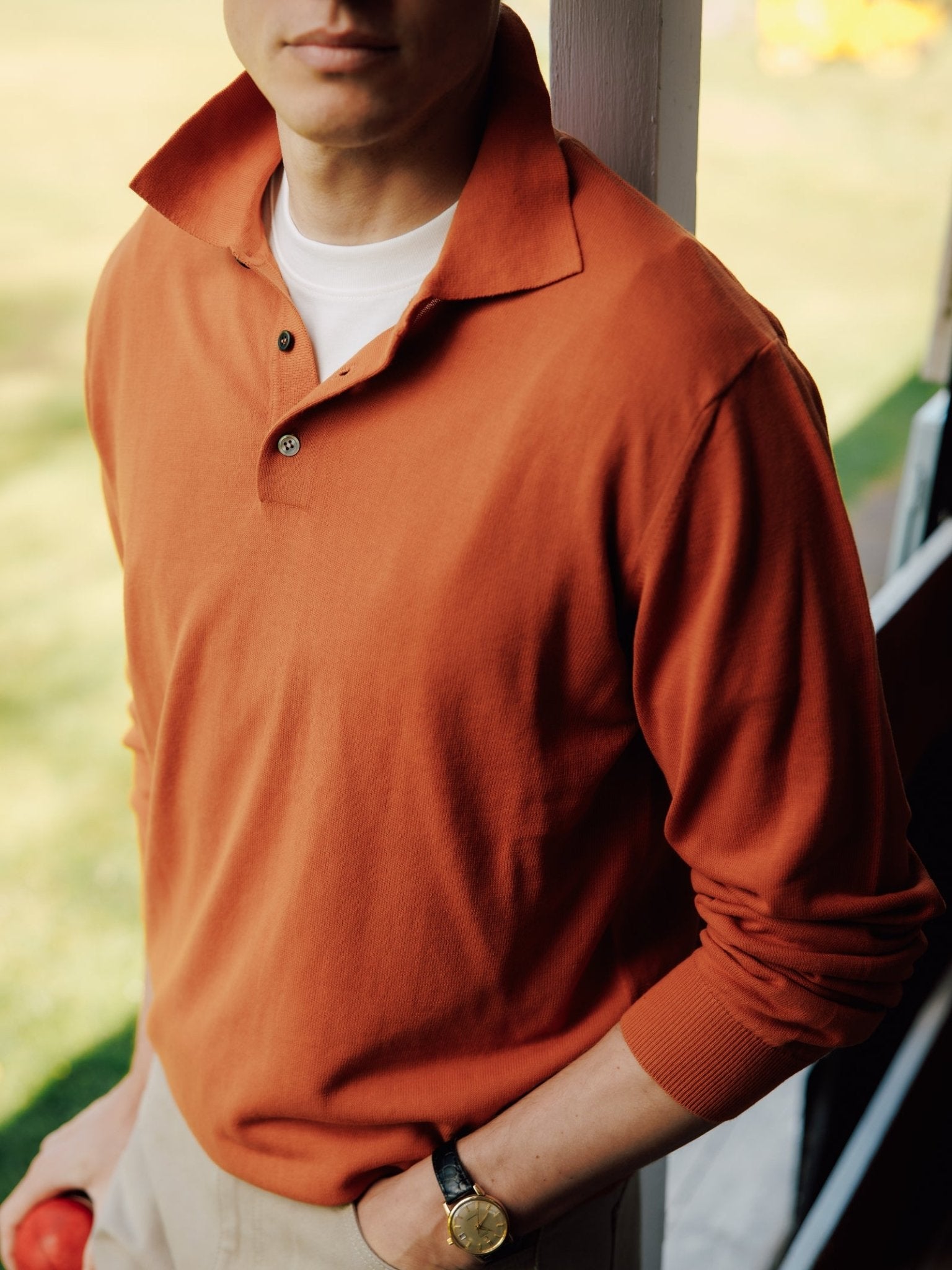 A person in a Campbell's of Beauly Cotton Rugby stands by a porch railing, holding an apple, with their other hand—wearing a watch—in their pocket. They're layering it over a white tee; their face is partly out of frame.