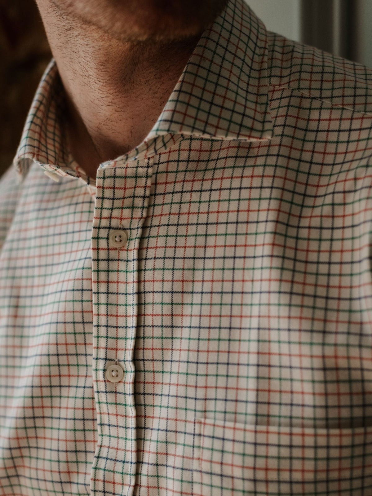 A close-up of a person wearing the Campbell’s of Beauly Windowpane Shirt, a beige brushed cotton button-up with a red, blue, and green checkered pattern. Only part of the neck and upper chest are visible.