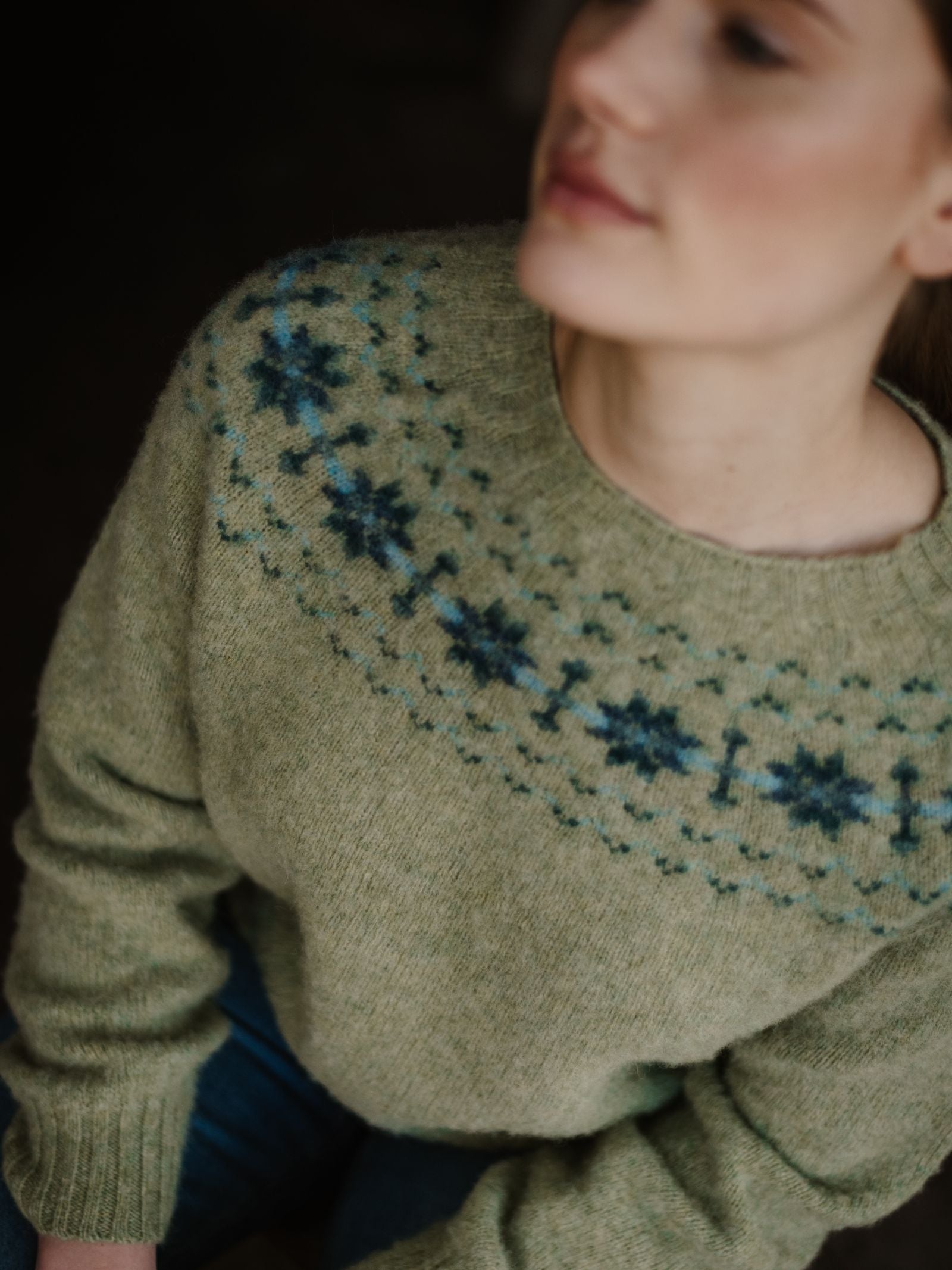 A woman sits looking away, her face partially blurred, wearing the Shaggy Cropped Fairisle Crew Jumper by Campbell's of Beauly—a light green knit featuring blue and dark green floral patterns around the neckline.